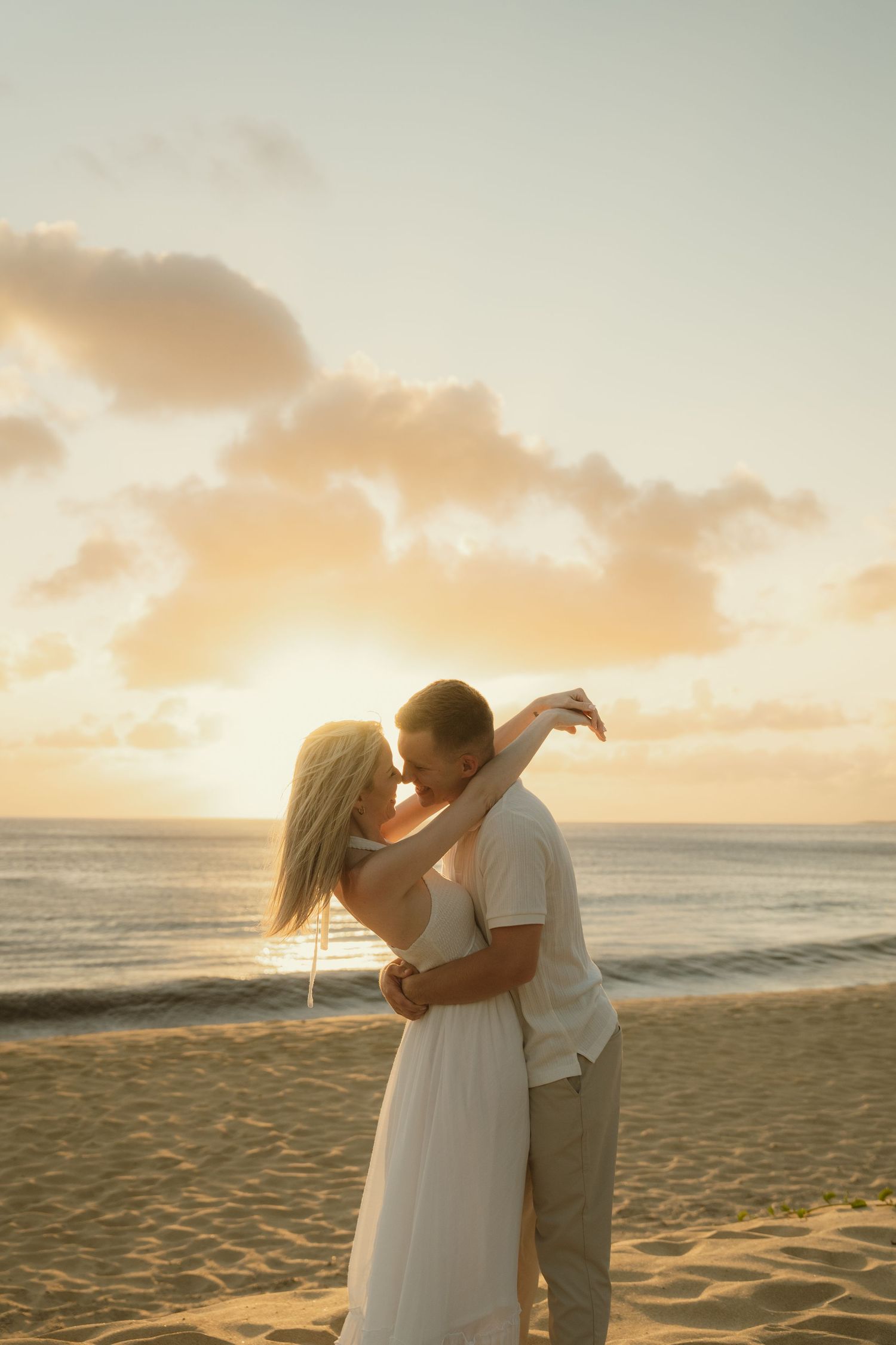 Couple sharing a romantic kiss on a sandy beach at golden sunset, woman in white dress, ocean waves in background.