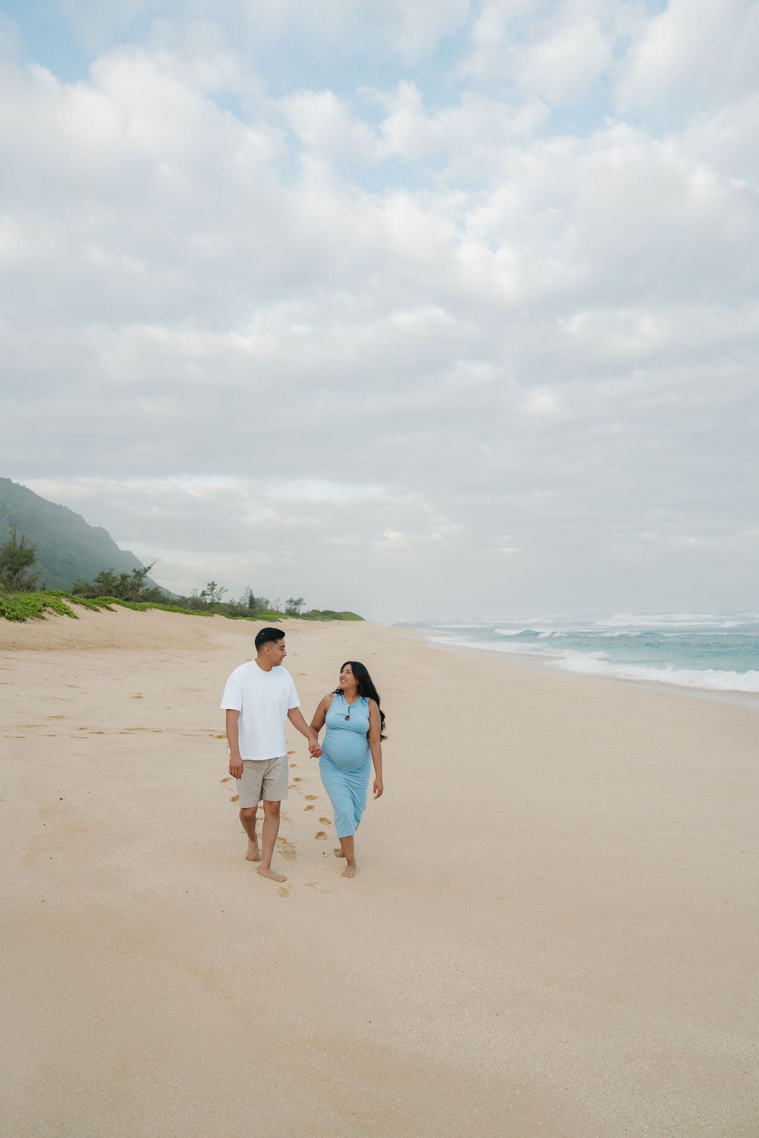 Expectant couple holding hands walking barefoot on a sandy beach together.
