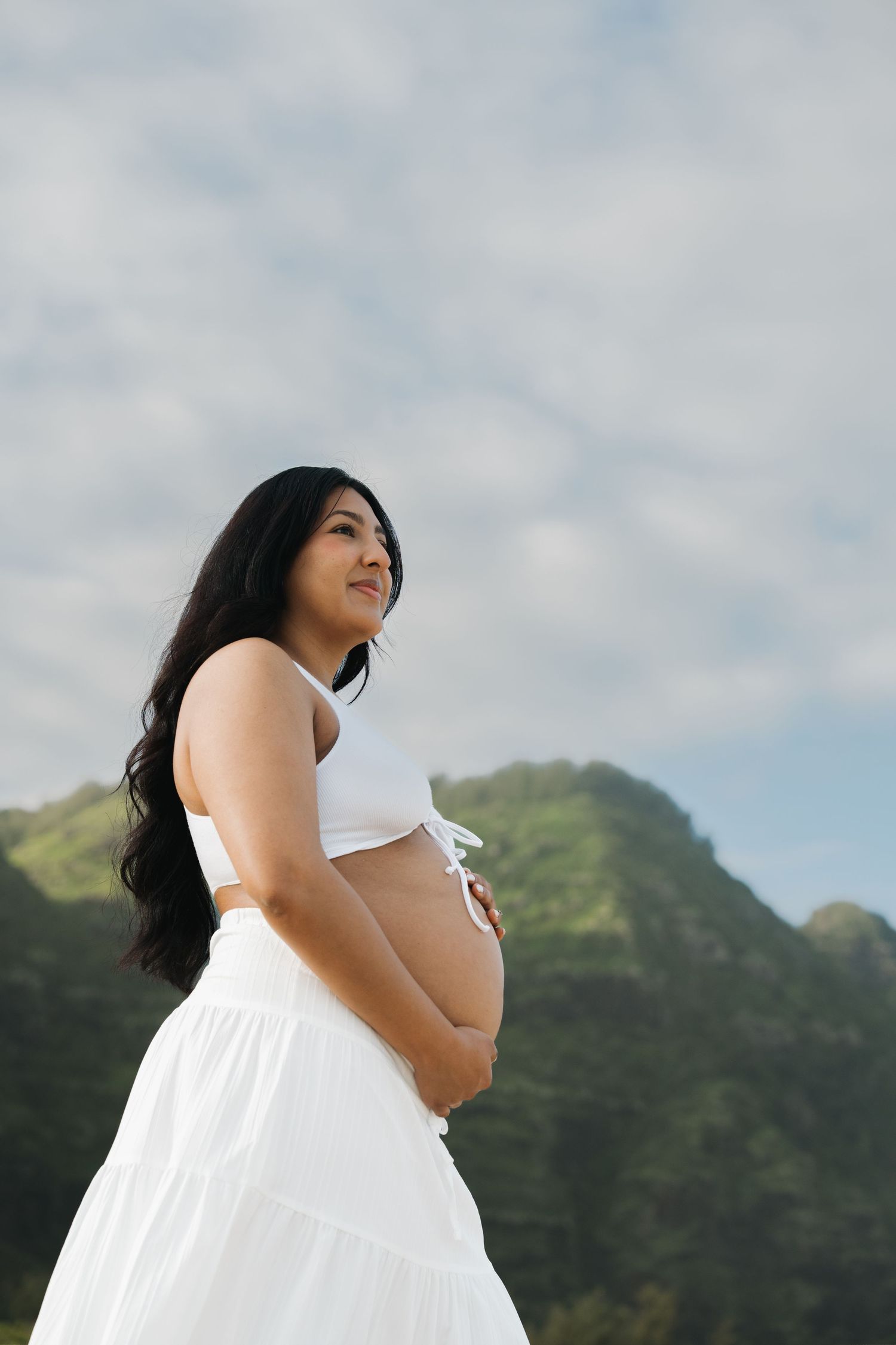 Smiling pregnant woman in white crop top and skirt cradling belly near mountains.