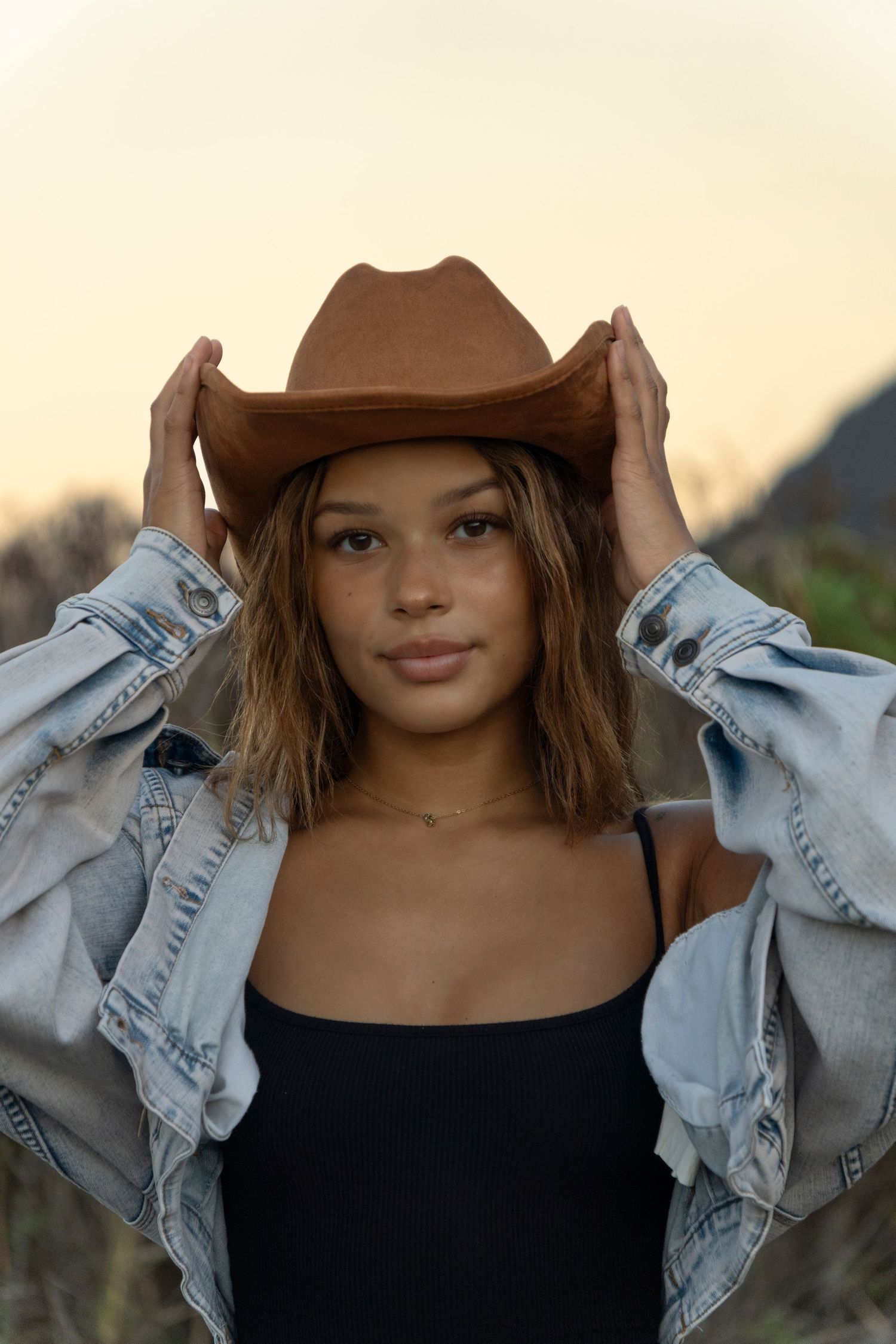 Young woman adjusting brown cowboy hat wearing denim jacket and black top at golden hour outdoors.