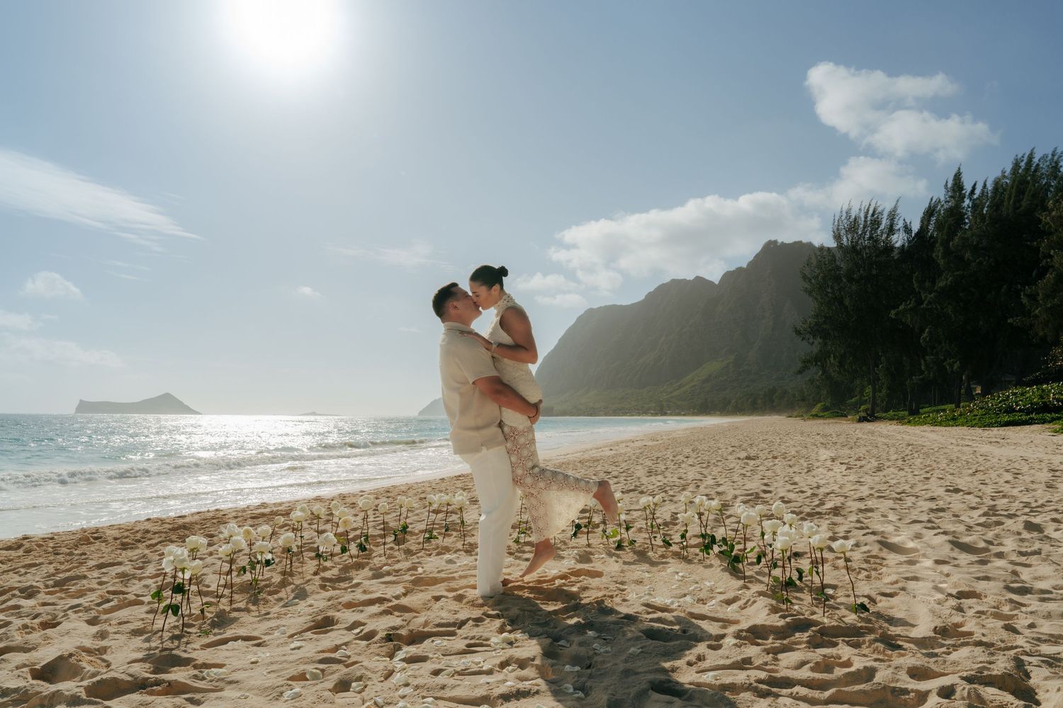 Couple sharing a romantic kiss on a sunny Hawaiian beach with dramatic mountain backdrop and golden sand.