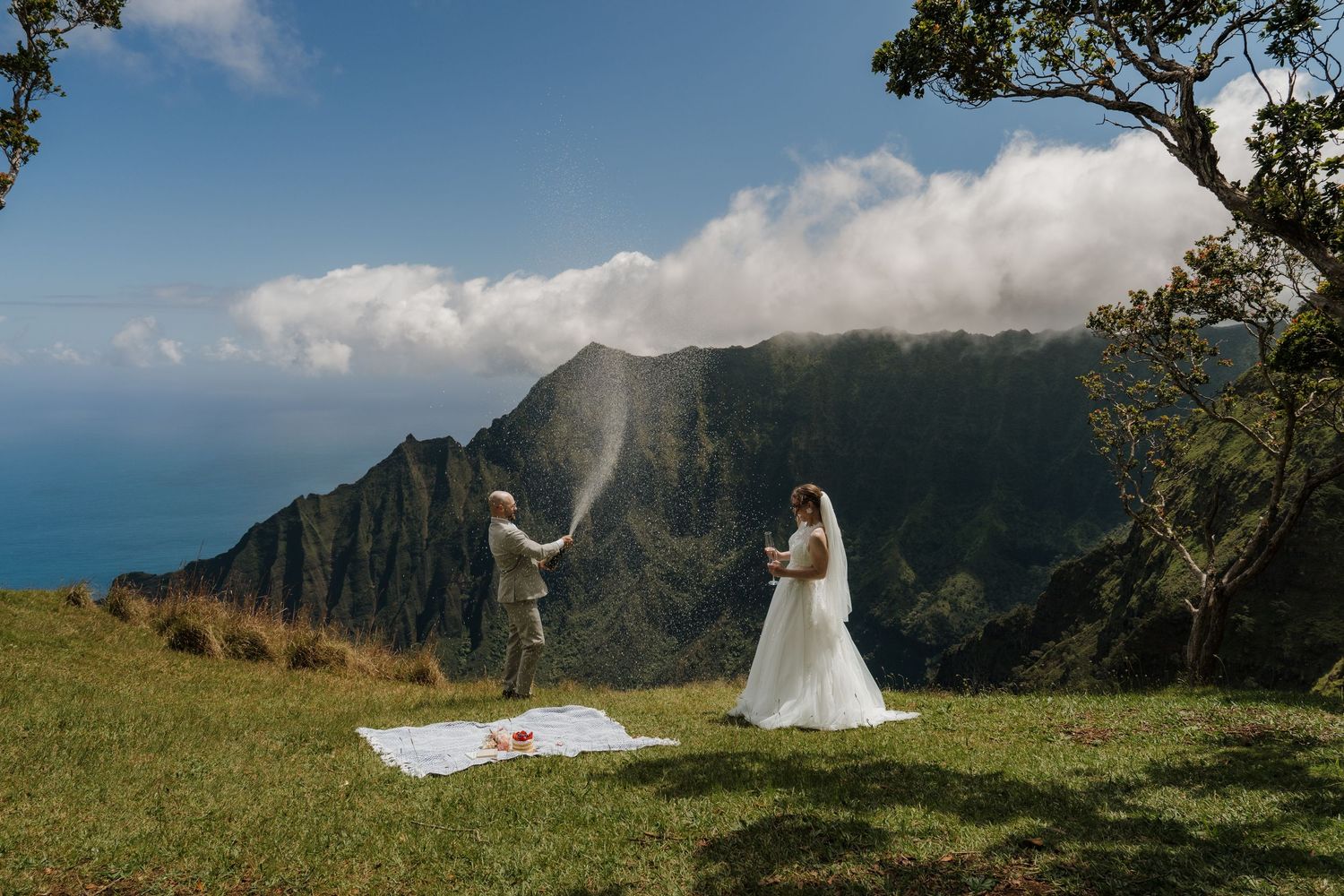 Groom spraying champagne celebrating with bride on green hillside overlooking Na Pali Coast Hawaii.