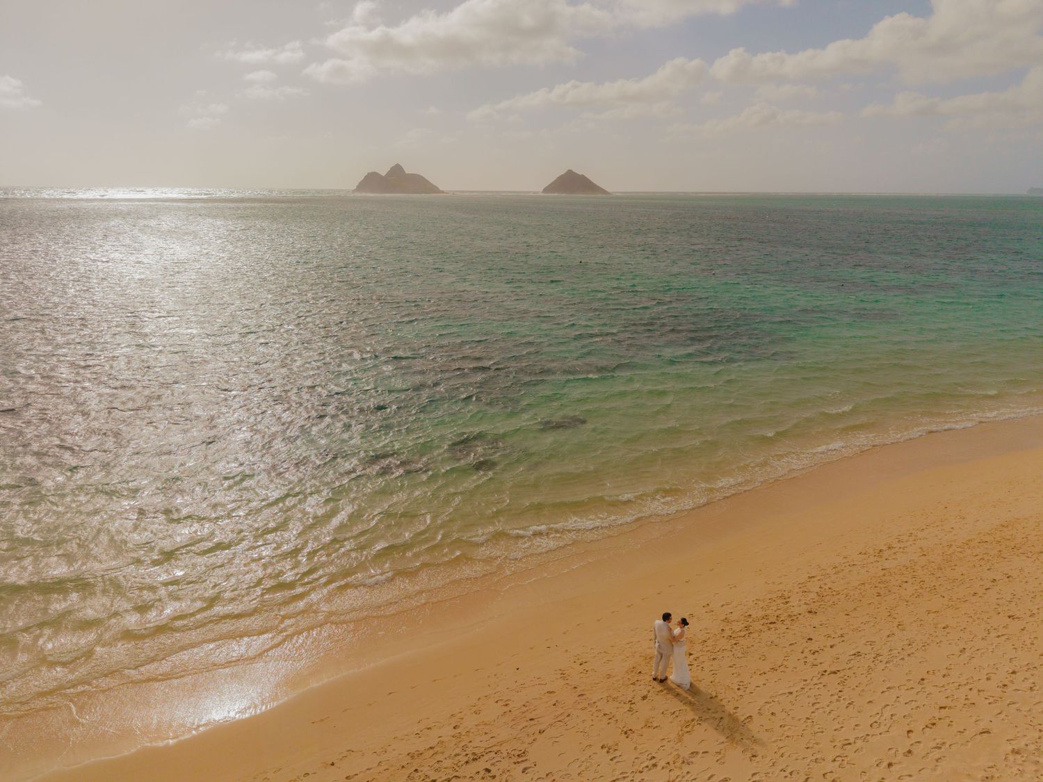Aerial view of a couple standing on a golden sandy beach with turquoise waters and two small islands in the distance.