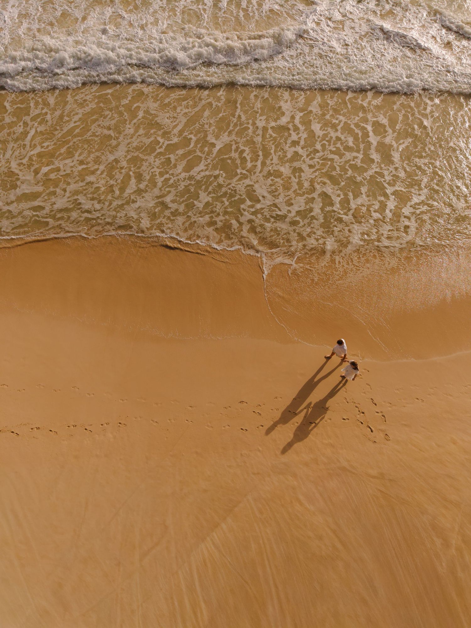 Aerial drone view of couple walking hand in hand along sandy beach near ocean waves.