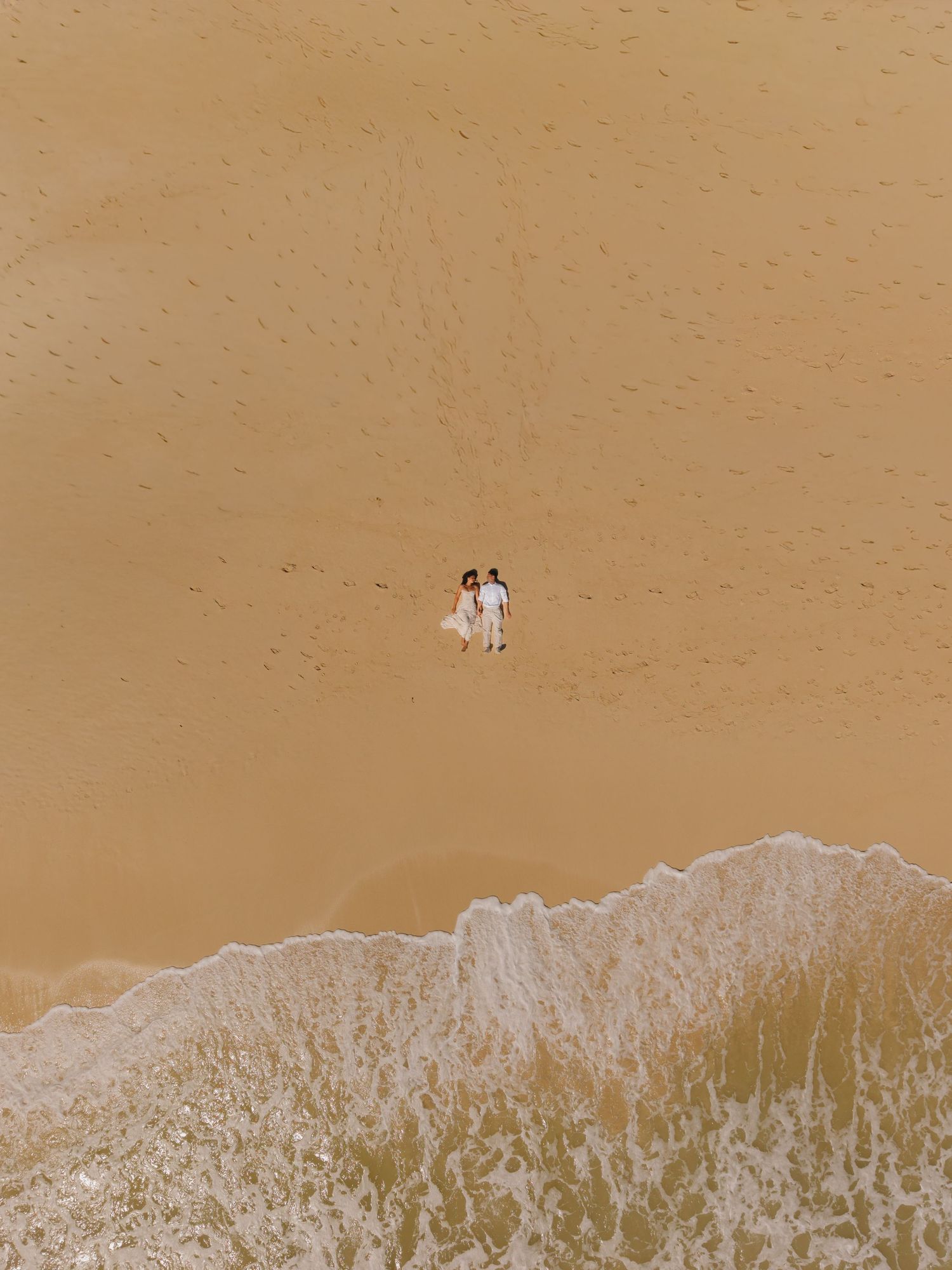 Aerial drone view of a couple in white standing on a sandy beach near ocean waves. drone kauai