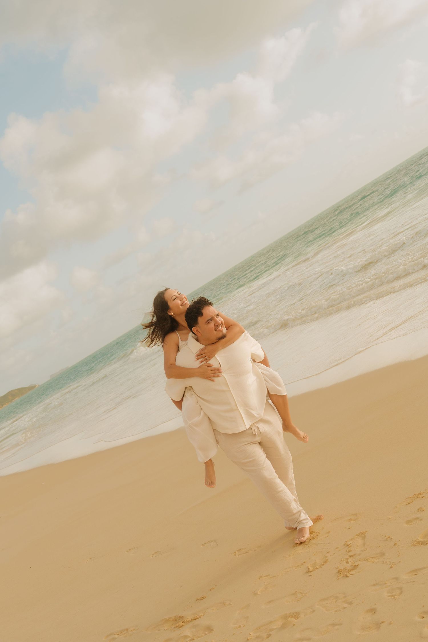 A happy couple enjoys a piggyback ride on a sandy beach with ocean waves and a cloudy sky behind them.