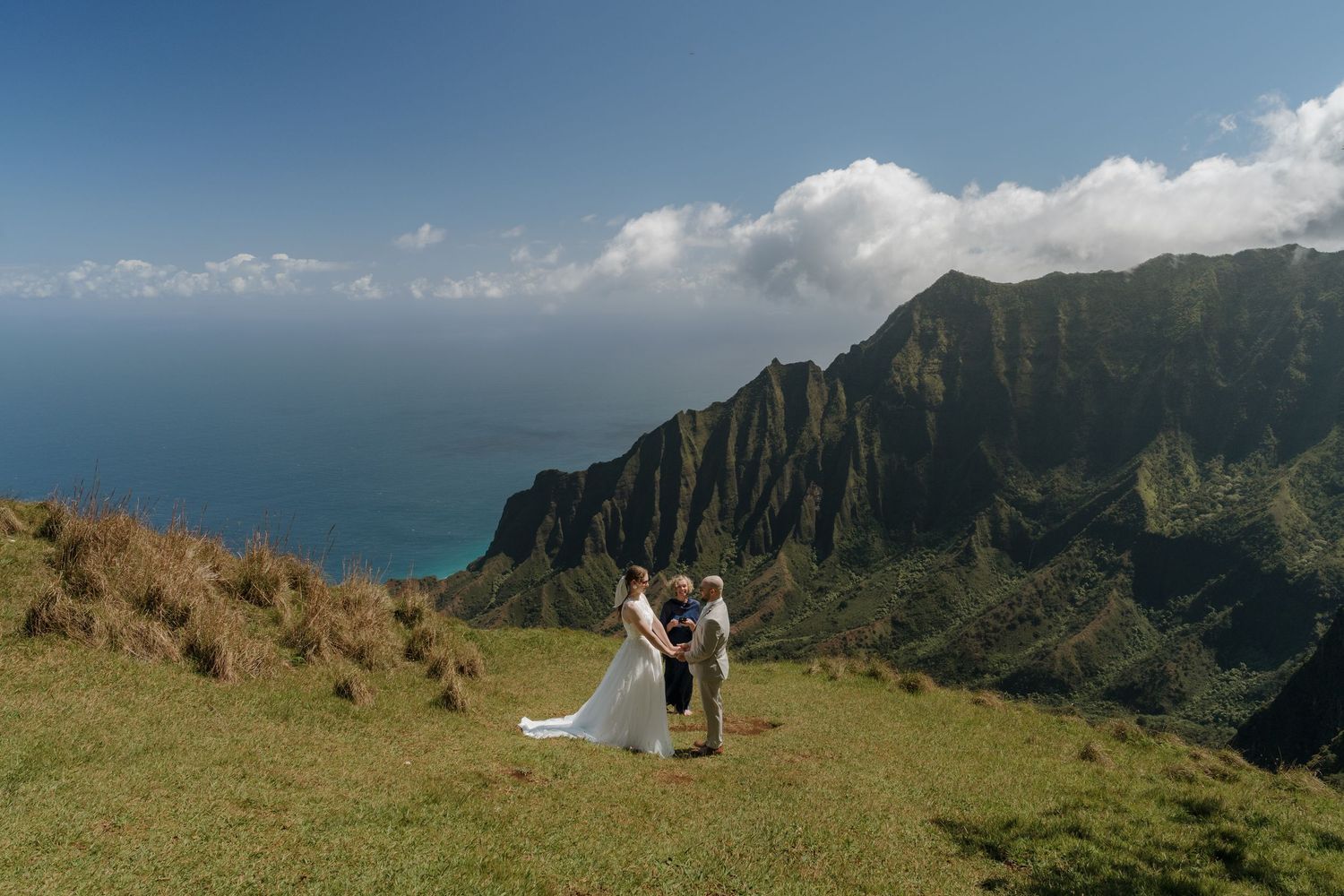 Couple exchanging vows on scenic Kauai mountaintop overlooking Na Pali Coast cliffs and ocean.