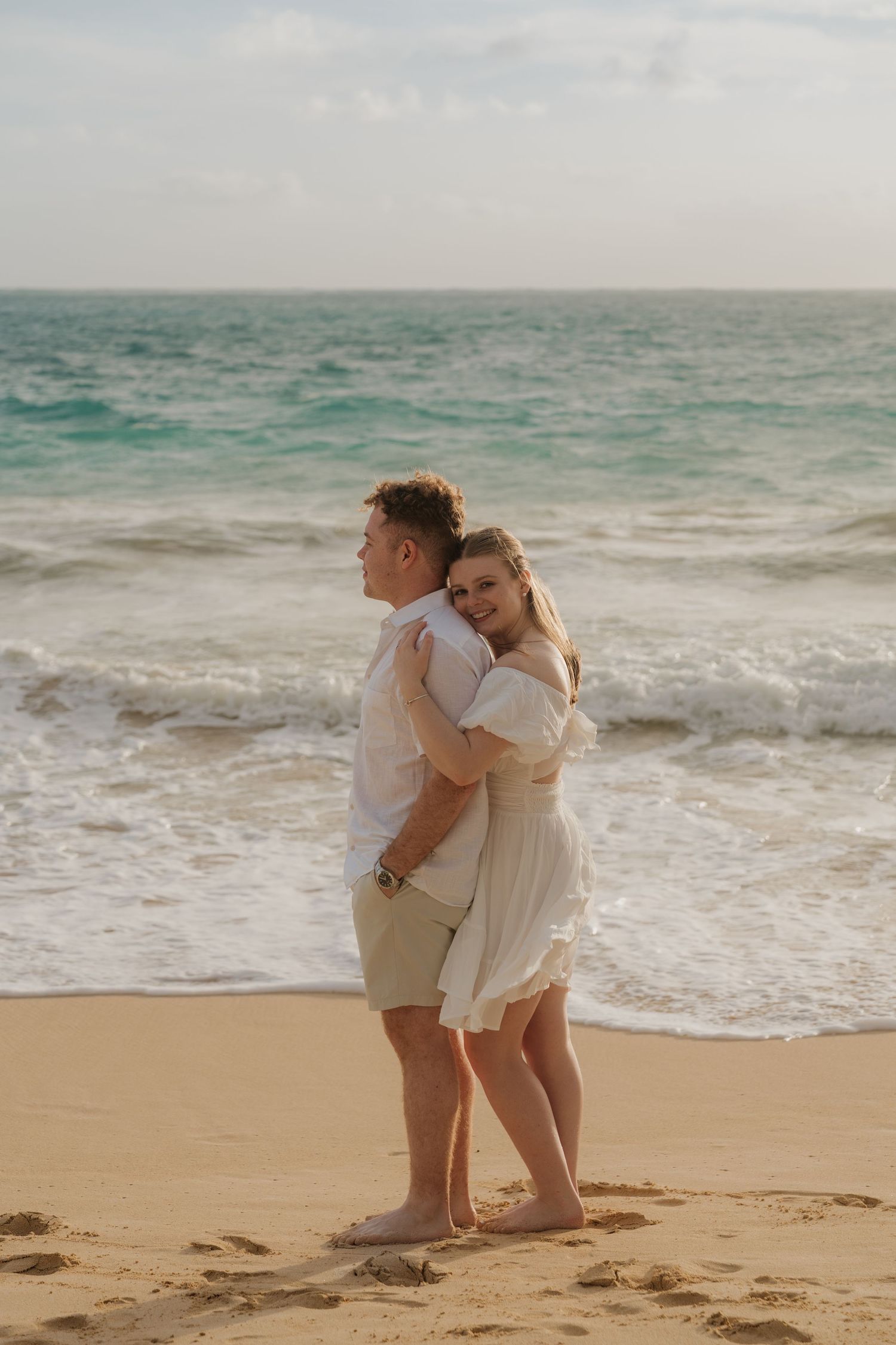 Couple embracing on a sandy beach with turquoise ocean waves, woman in white dress smiling at camera.