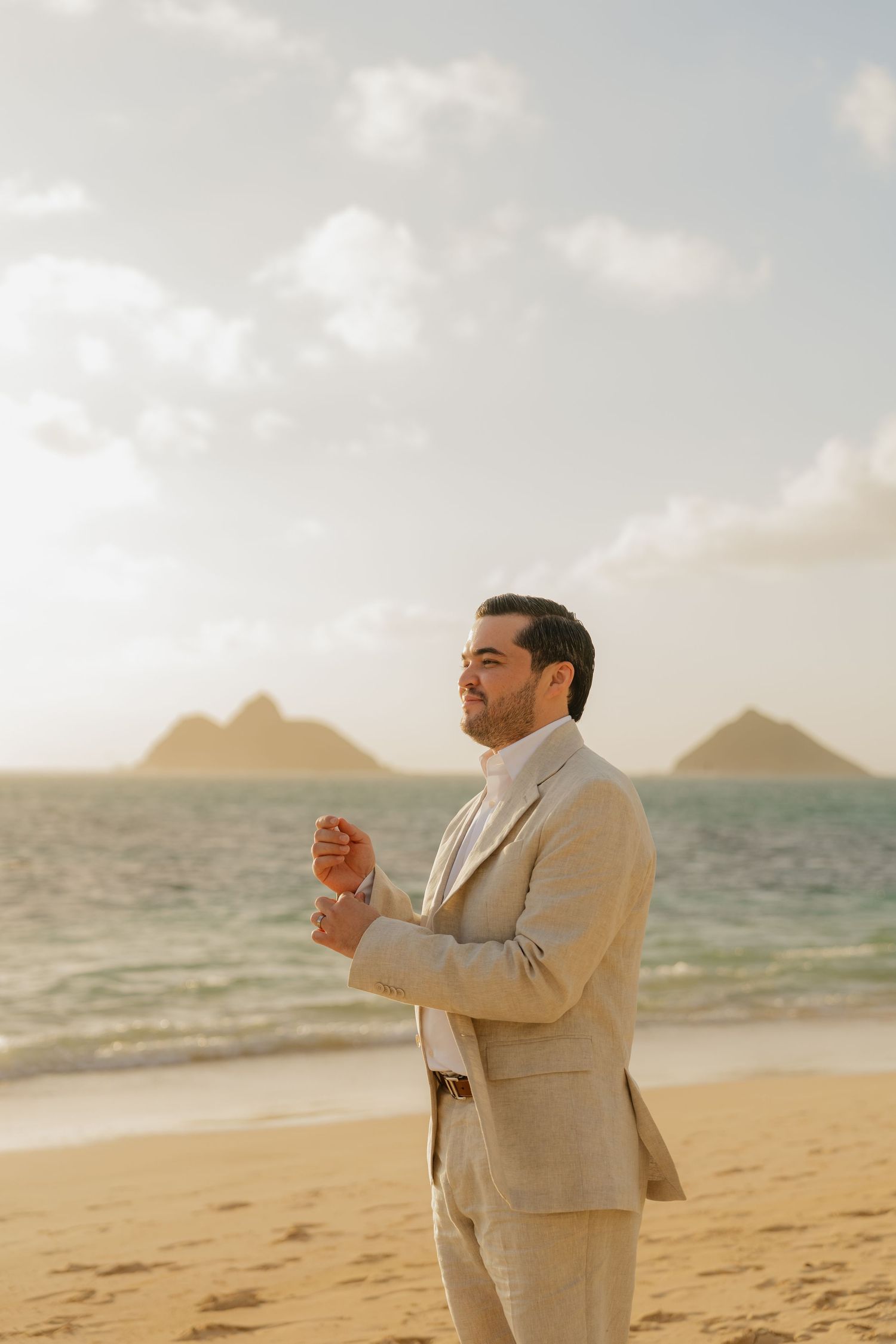 Groom in beige linen suit adjusting cufflinks on sandy beach with twin island peaks and ocean backdrop.