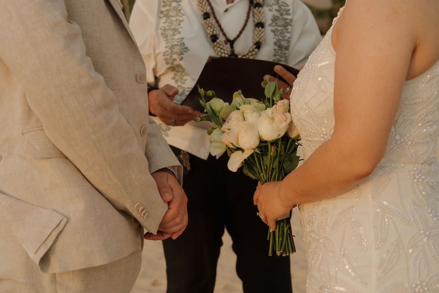 Bride holding white floral bouquet while exchanging vows with groom, officiant in traditional attire at wedding ceremony.