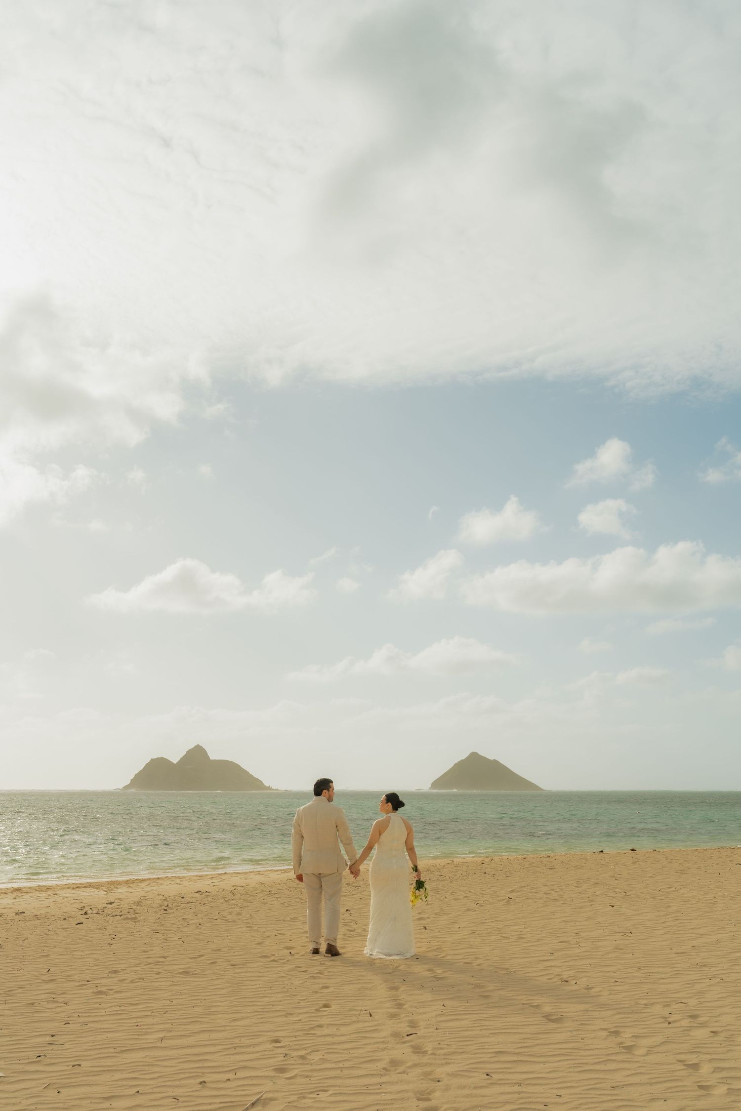 Bride and groom holding hands on sandy beach with twin island peaks and blue ocean backdrop in Hawaii.