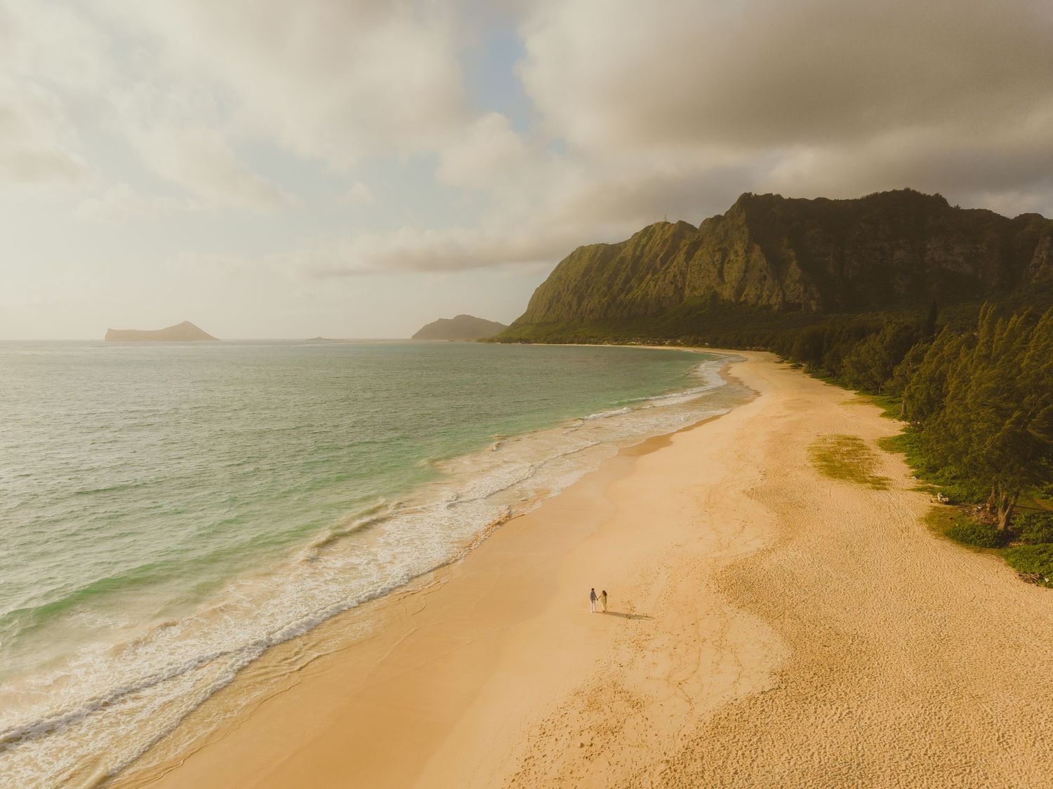 Aerial view of a pristine sandy beach in Hawaii with turquoise waves, lush green mountains, and two people walking along the shore.