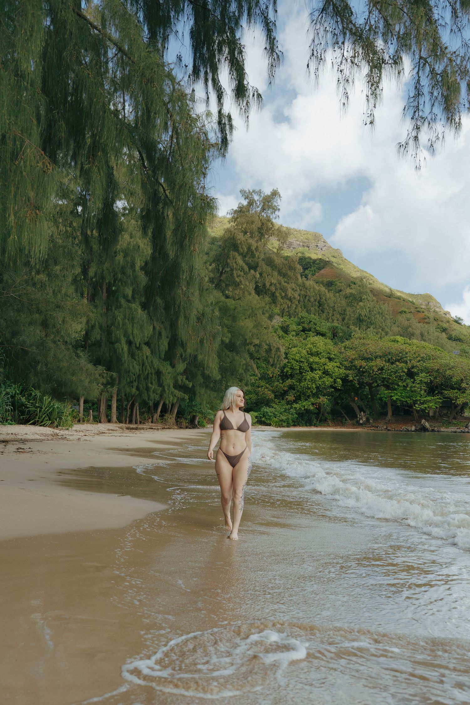 Woman in brown bikini walking along sandy tropical beach with lush green mountains.