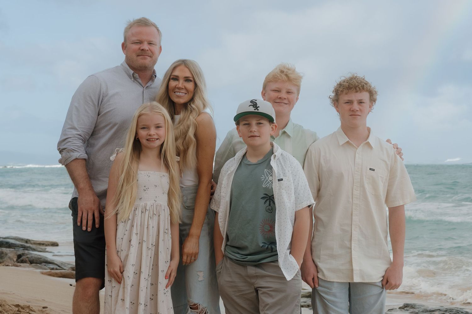 Happy family of six posing together on a tropical beach with ocean waves behind them.