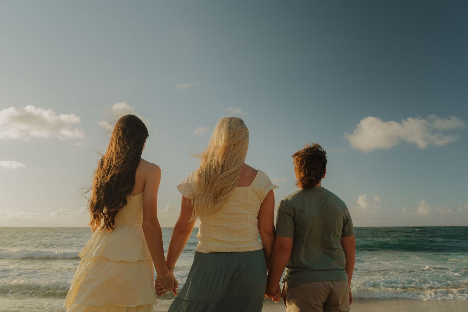 Three friends holding hands on a tropical beach, viewed from behind, facing the ocean under a blue sky.