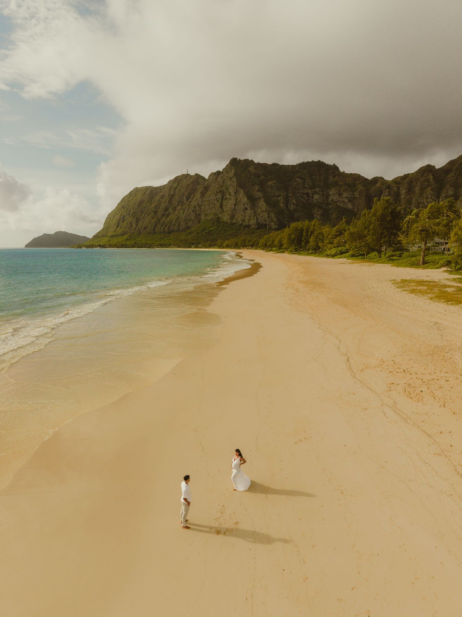 Aerial view of a couple in white attire on a golden sandy beach in Hawaii with turquoise water and lush mountains.
