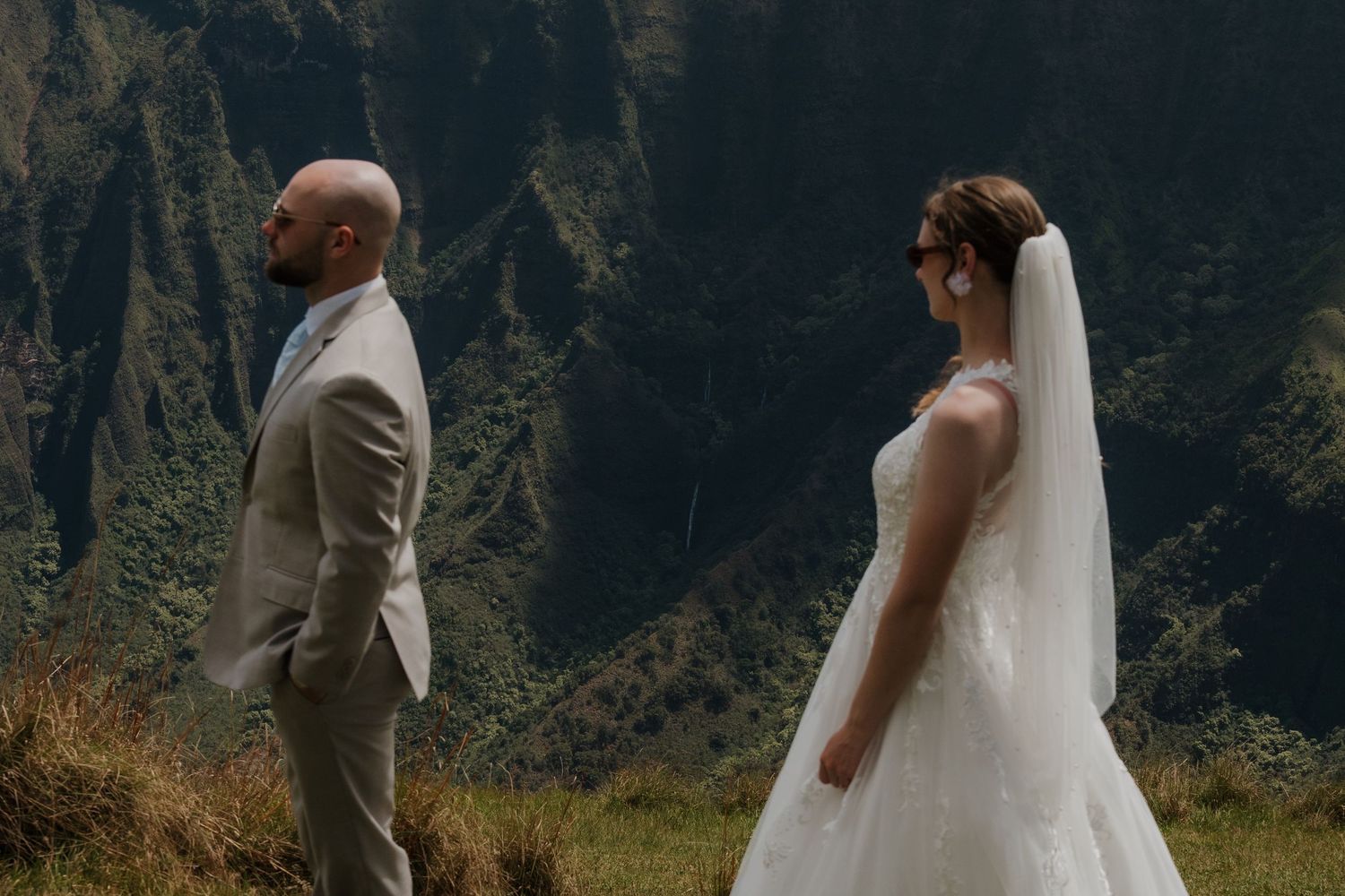 Bride in white gown and veil stands facing groom in tan suit during outdoor mountain wedding ceremony in Hawaii.