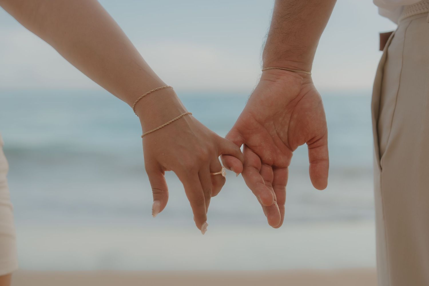 Close-up of couple gently touching fingers with engagement ring on beach.