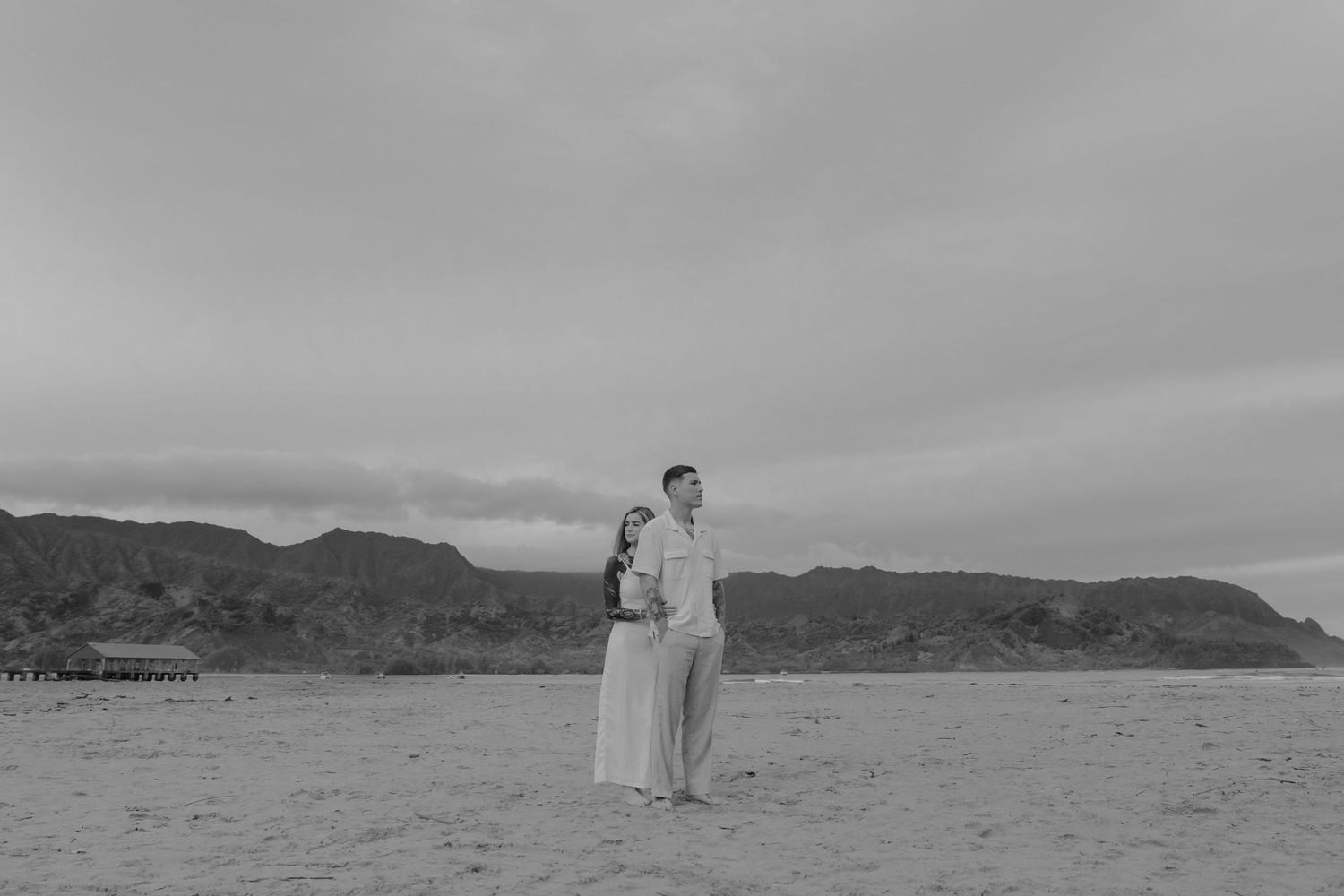 Black and white photo of couple standing back to back on vast beach with mountain backdrop.