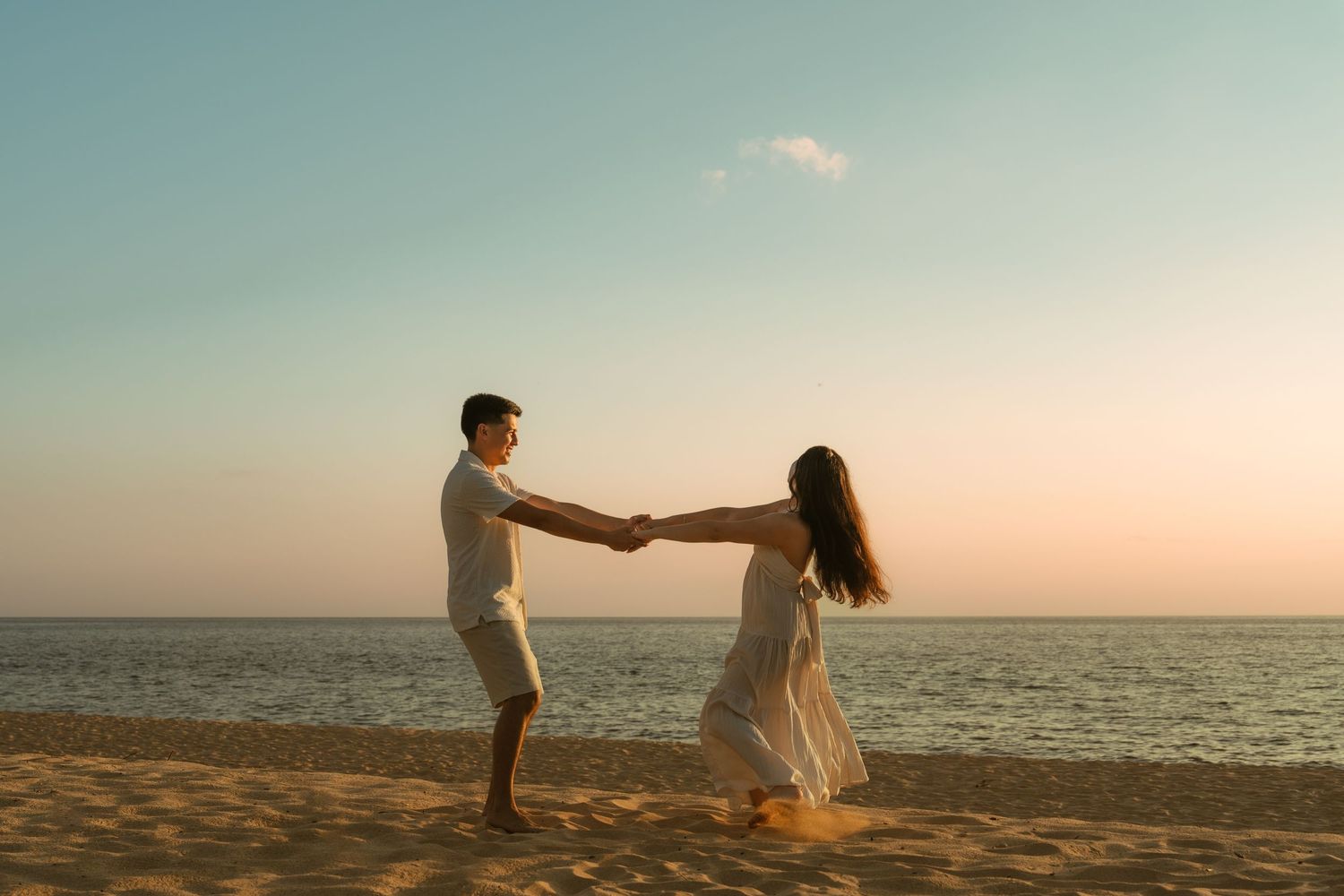 Couple dancing and holding hands on a sandy beach at sunset with calm ocean waters in the background.