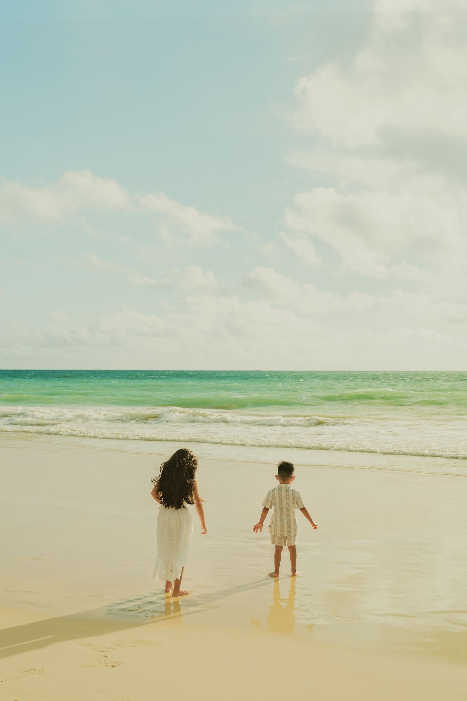 A woman and child walk barefoot on a sunny sandy beach toward turquoise ocean waves under a bright blue sky.