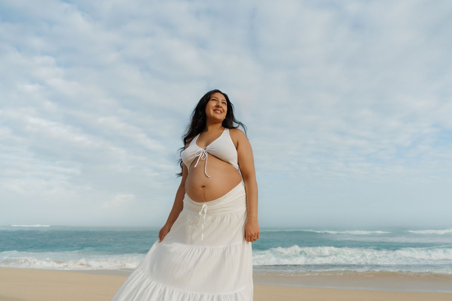 Pregnant woman in white crop top and skirt posing on tropical beach with ocean waves behind her.
