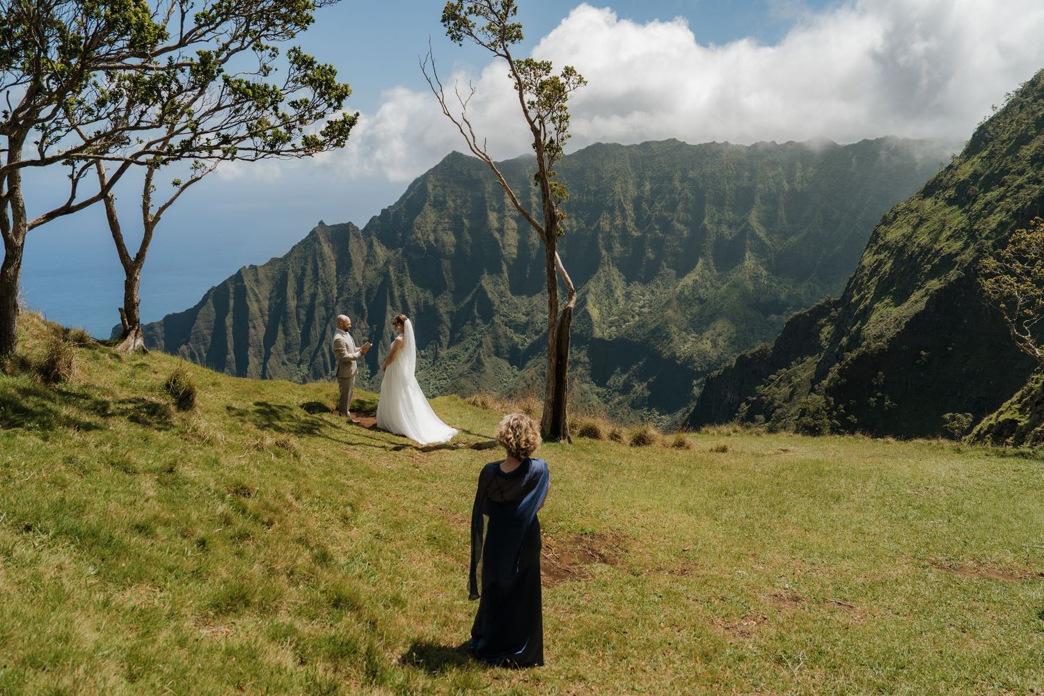 Bride in white gown and guest in black dress stand on lush green hillside with dramatic Kauai Na Pali Coast mountains backdrop.