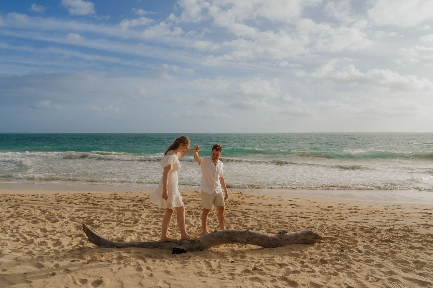 Two women in white dresses holding hands near driftwood on a sunny tropical beach.