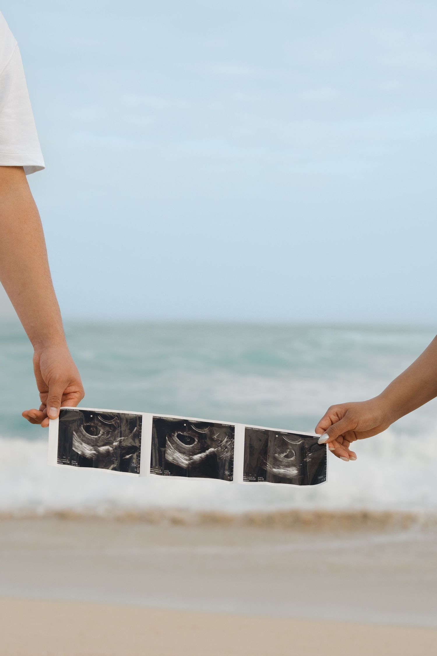 Couple holding ultrasound photos together on a beach with ocean backdrop.