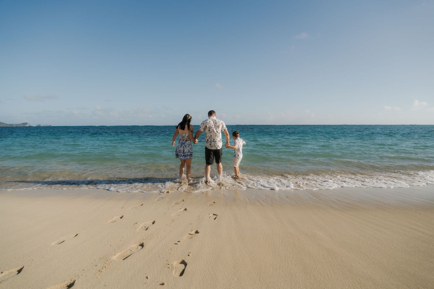 Family of three standing at the shoreline of a tropical beach gazing at the turquoise ocean.