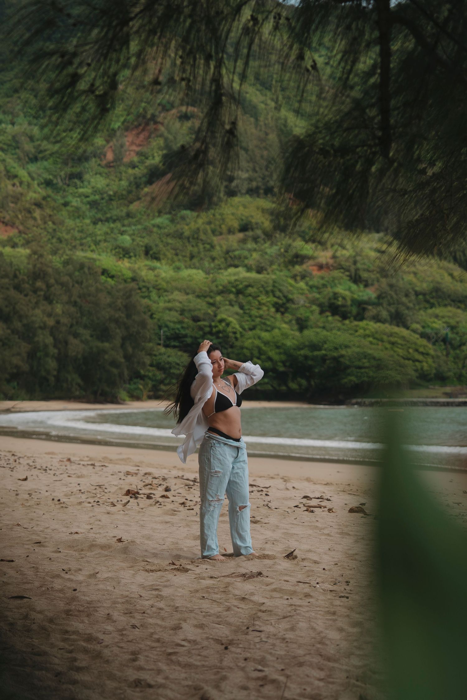 Woman in white shirt and jeans posing on tropical beach with lush green cliffs.