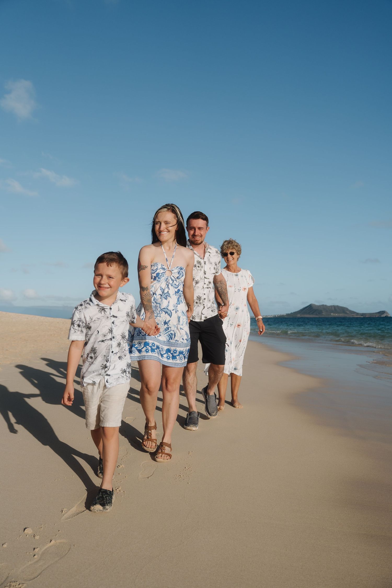 Family of four walking along a sunny Hawaiian beach with turquoise water and mountains in background.