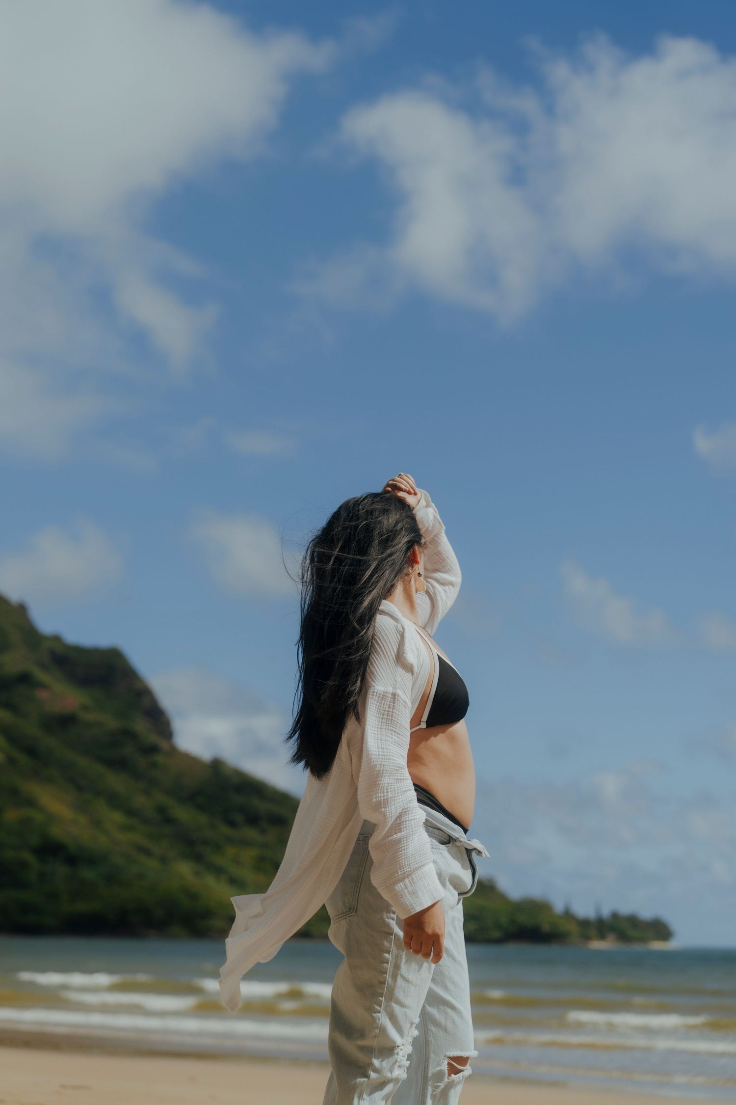 Woman in white shirt and jeans on sunny tropical beach with mountains in background.