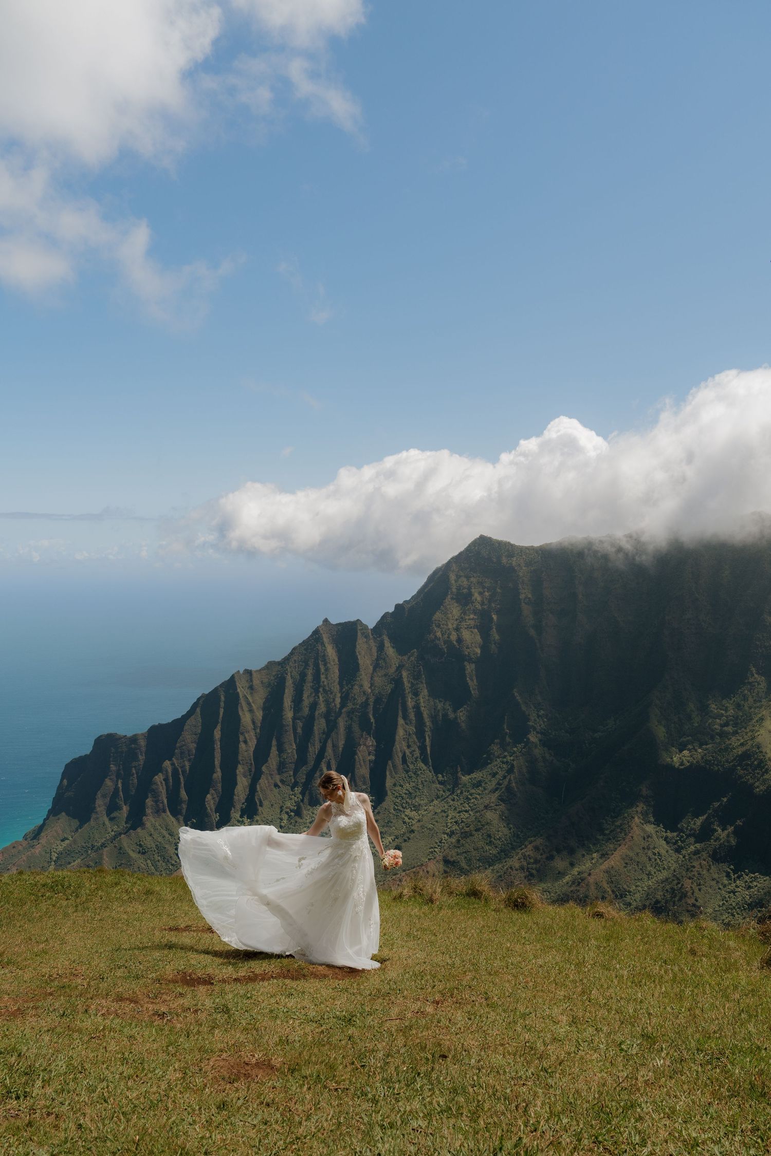 Bride in flowing white dress stands on green hillside with dramatic Na Pali Coast mountains and blue ocean in background.