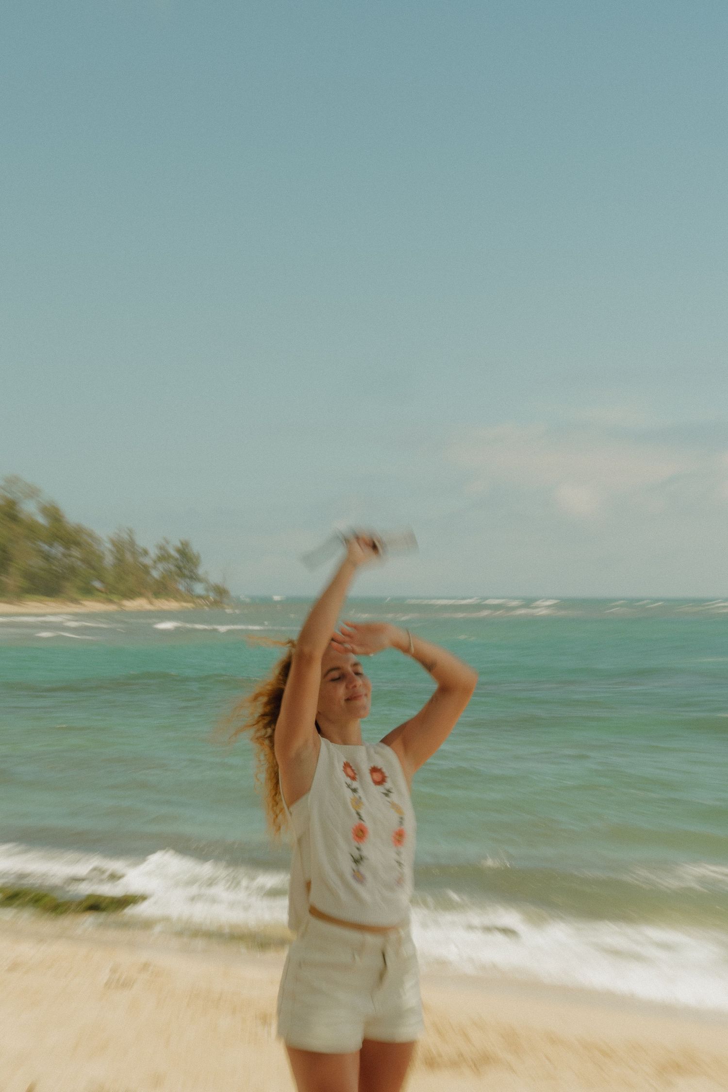 Blonde woman in white crop top and shorts posing on a tropical beach with turquoise ocean waves.