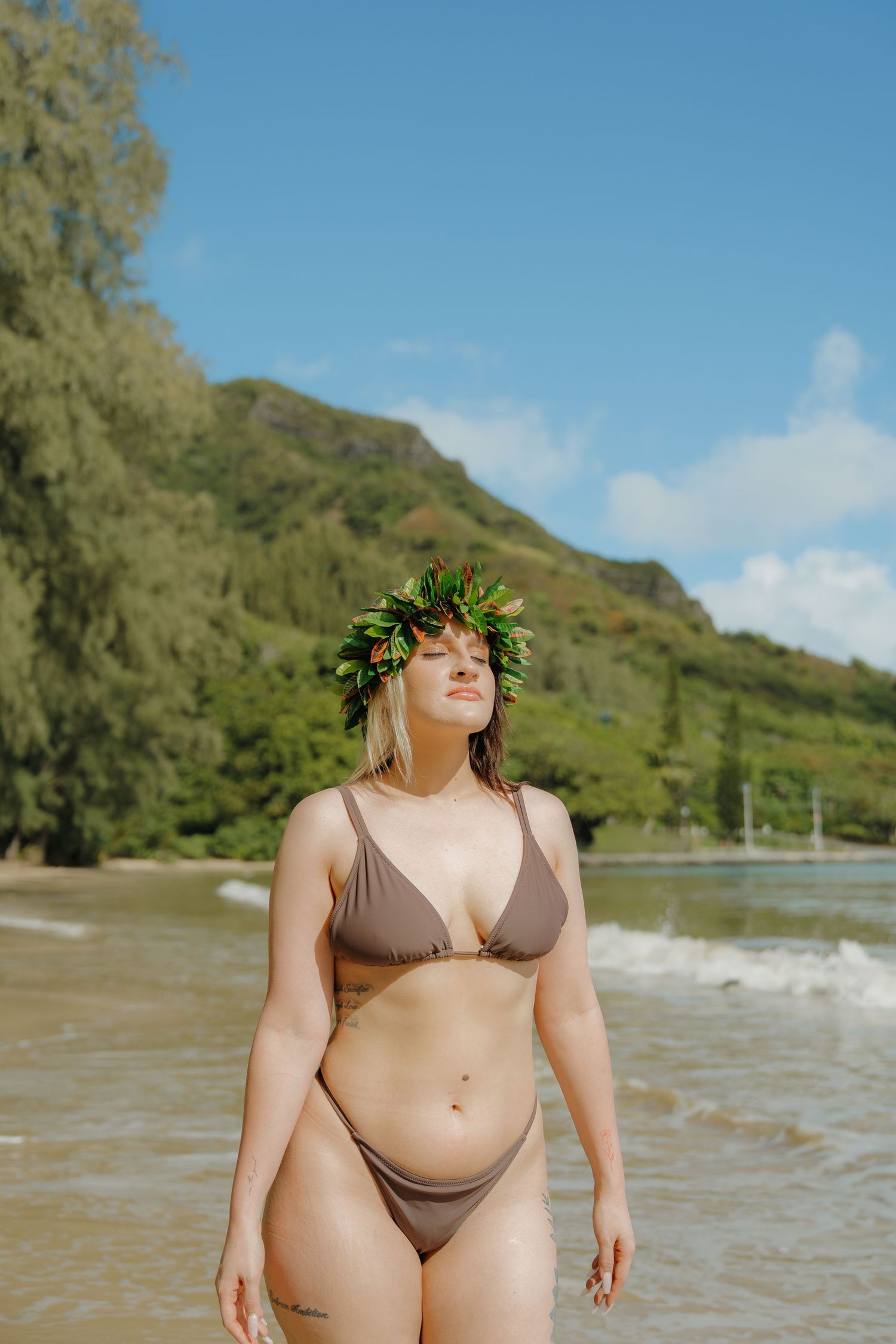Woman in brown bikini wearing tropical leaf crown standing on sunny Hawaiian beach.