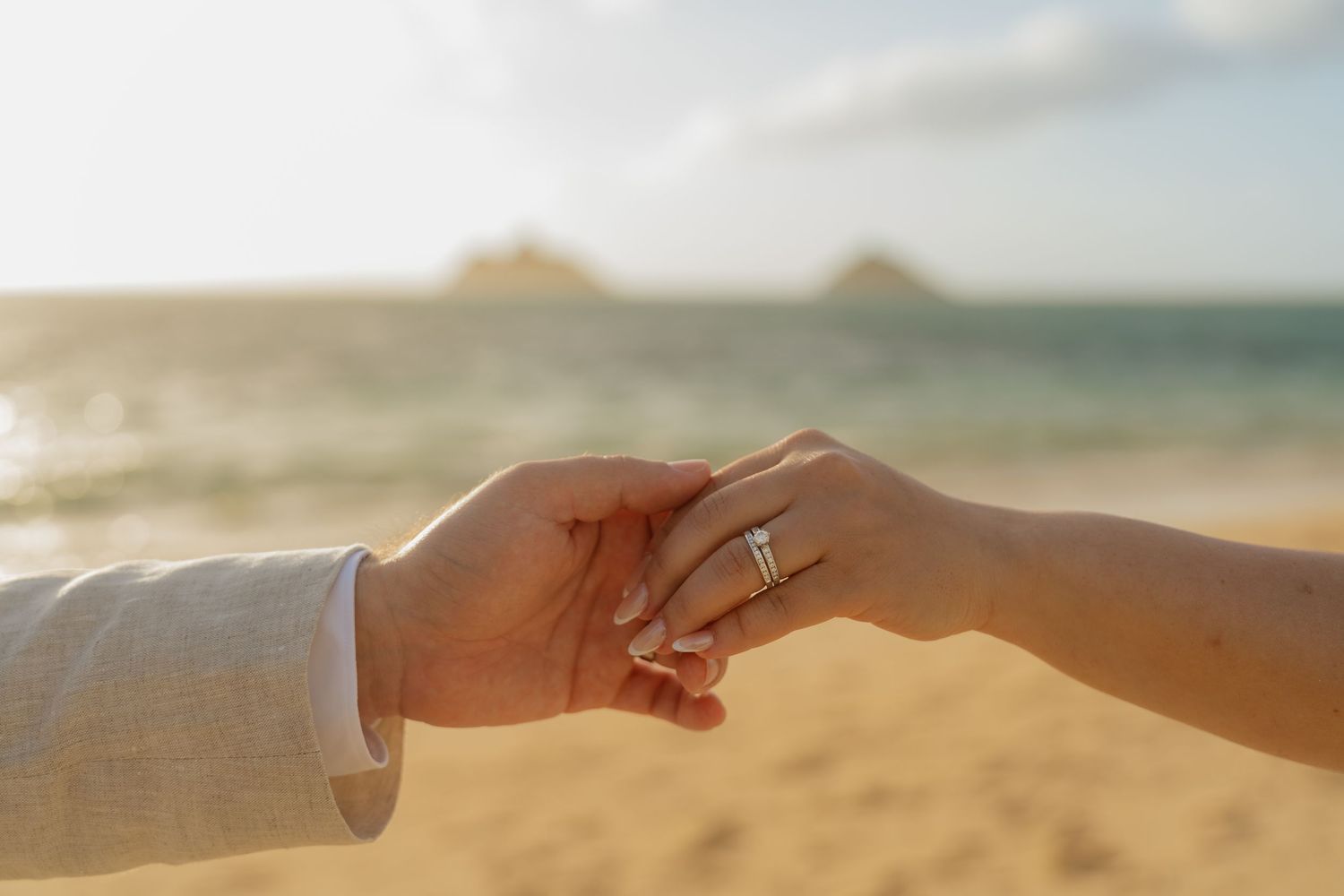 Couple holding hands on a beach at sunset, woman wearing a diamond engagement ring, ocean and islands in background.