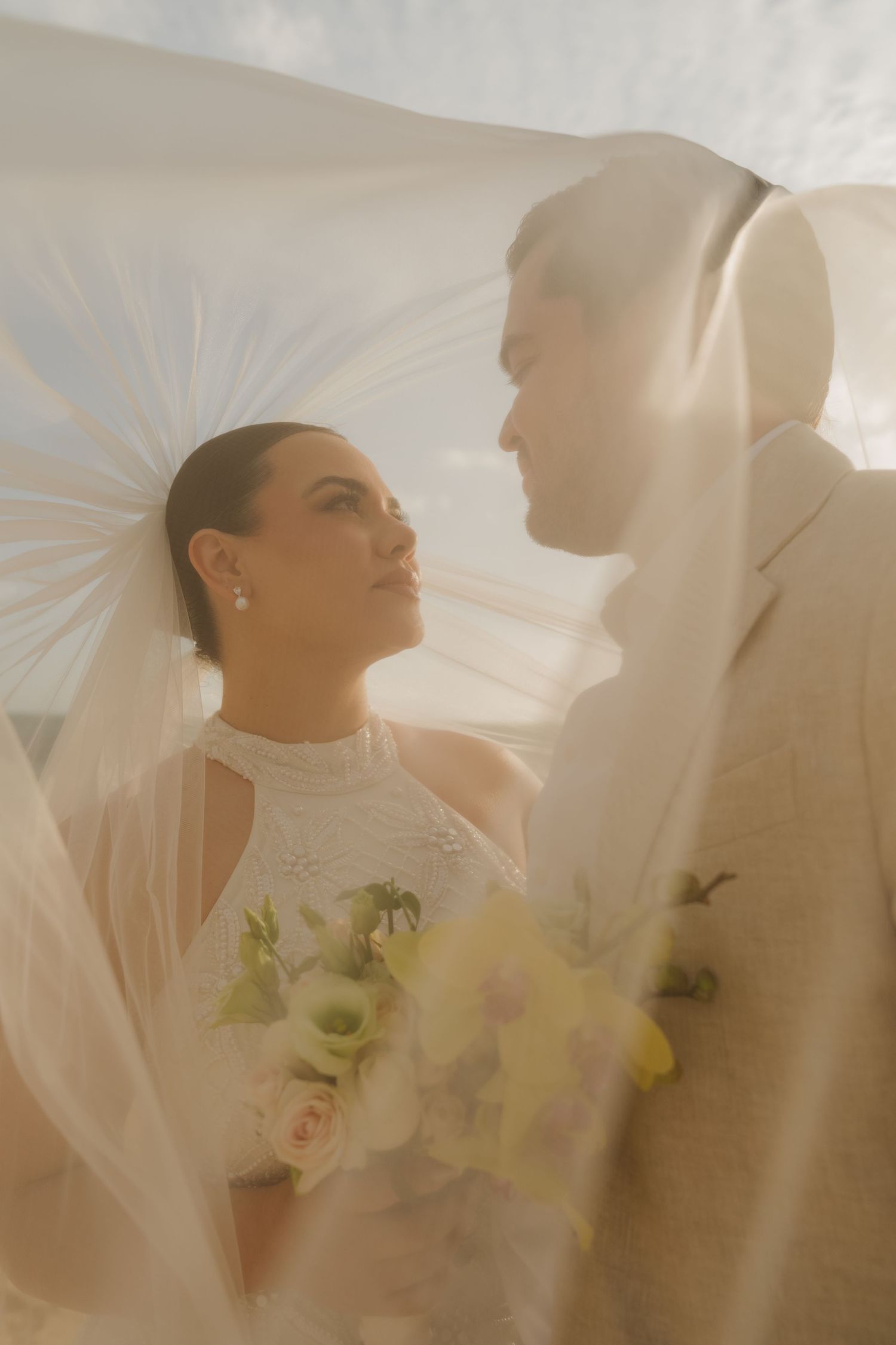 Bride and groom gazing at each other under a flowing white veil, bride holding a bouquet of white and yellow flowers.