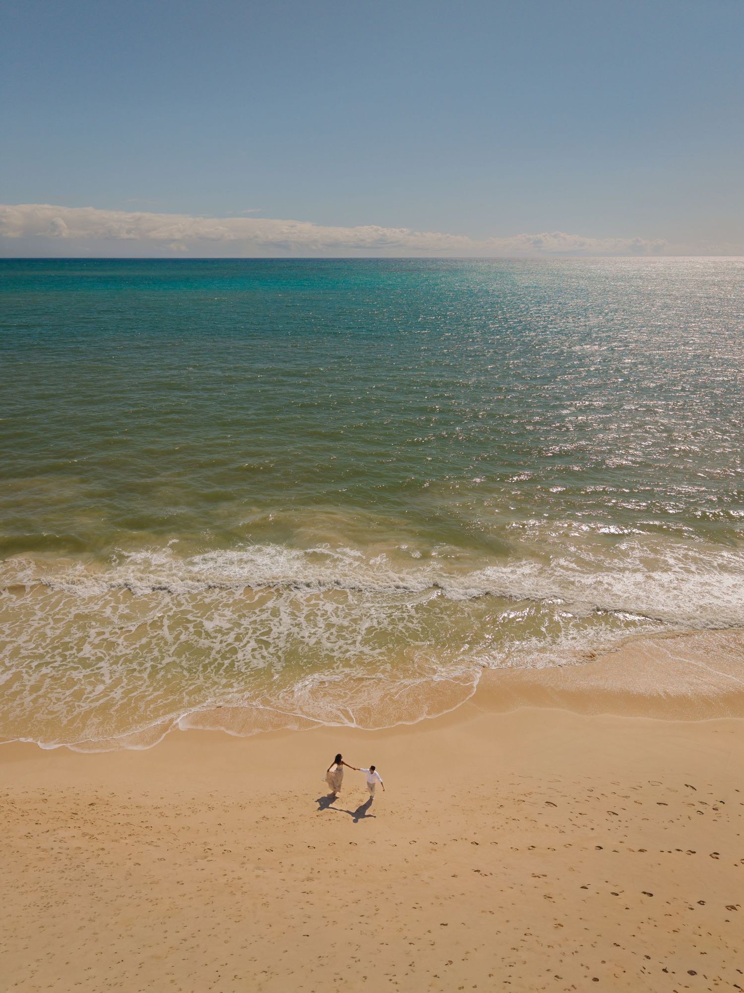 Aerial view of two people walking hand in hand on a sunny sandy beach with turquoise ocean waves. drone Hawaii Oahu