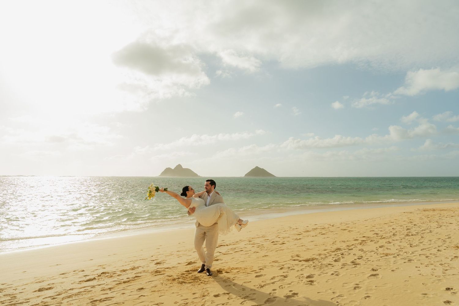 Groom lifting bride on sandy Hawaiian beach with turquoise ocean and Mokulua Islands in background.
