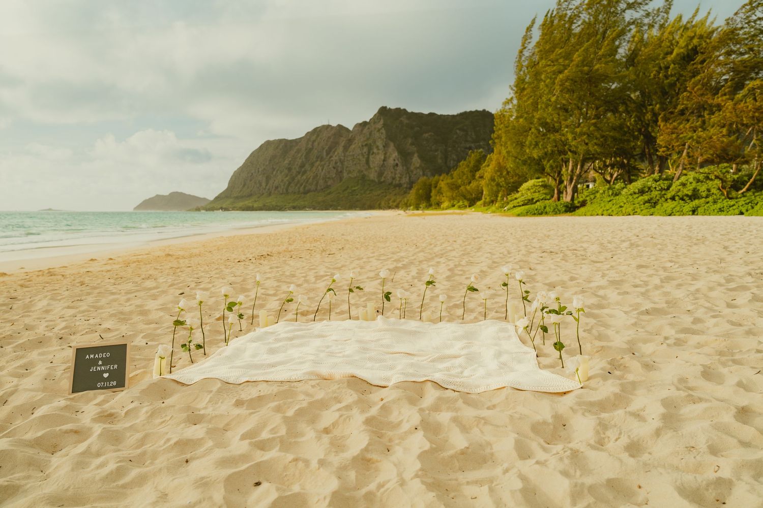 Romantic beach wedding setup with white floral arrangement on sand, framed by lush mountains in Hawaii.