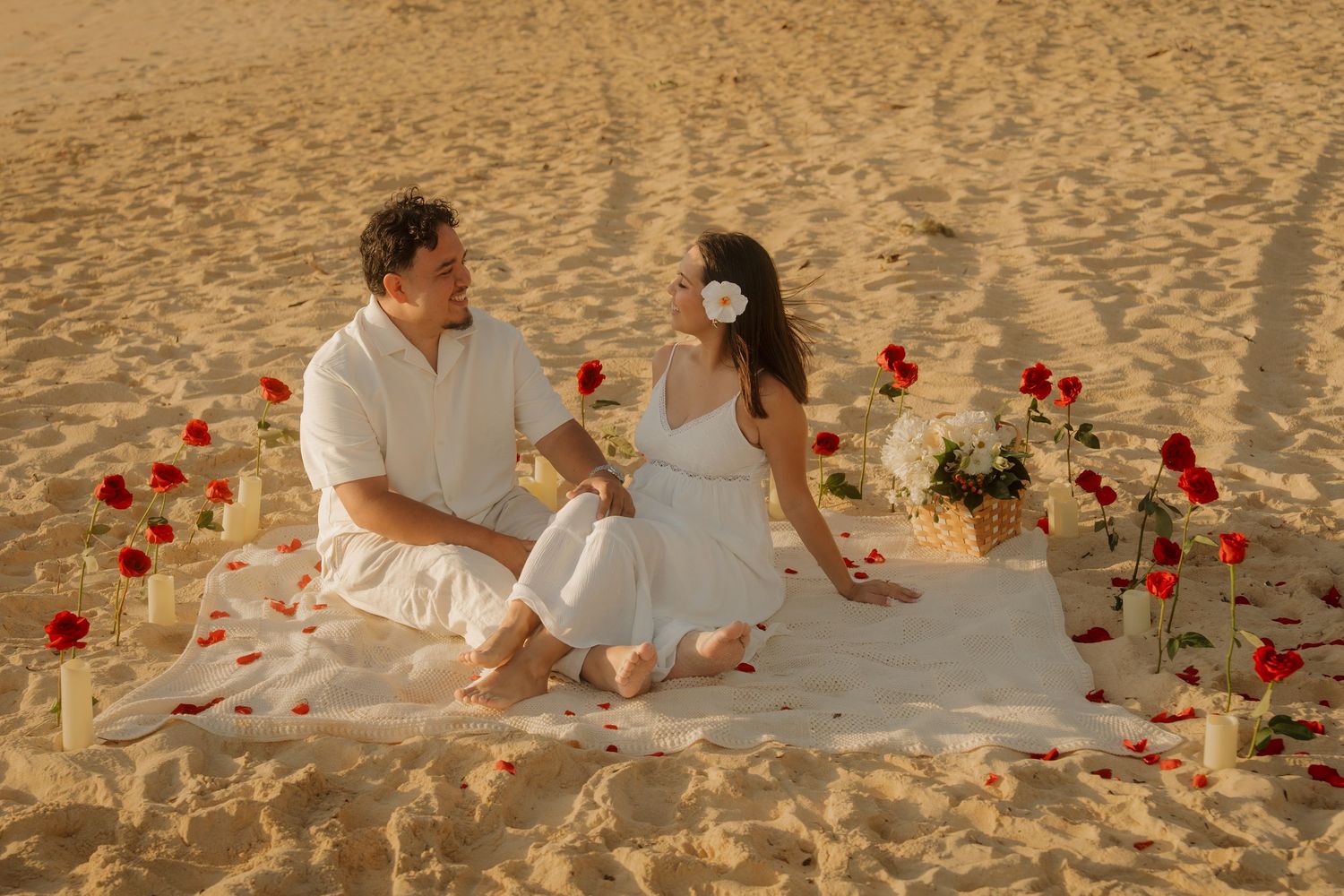 Romantic couple in white outfits sitting on a blanket in sandy desert surrounded by red roses and candles.