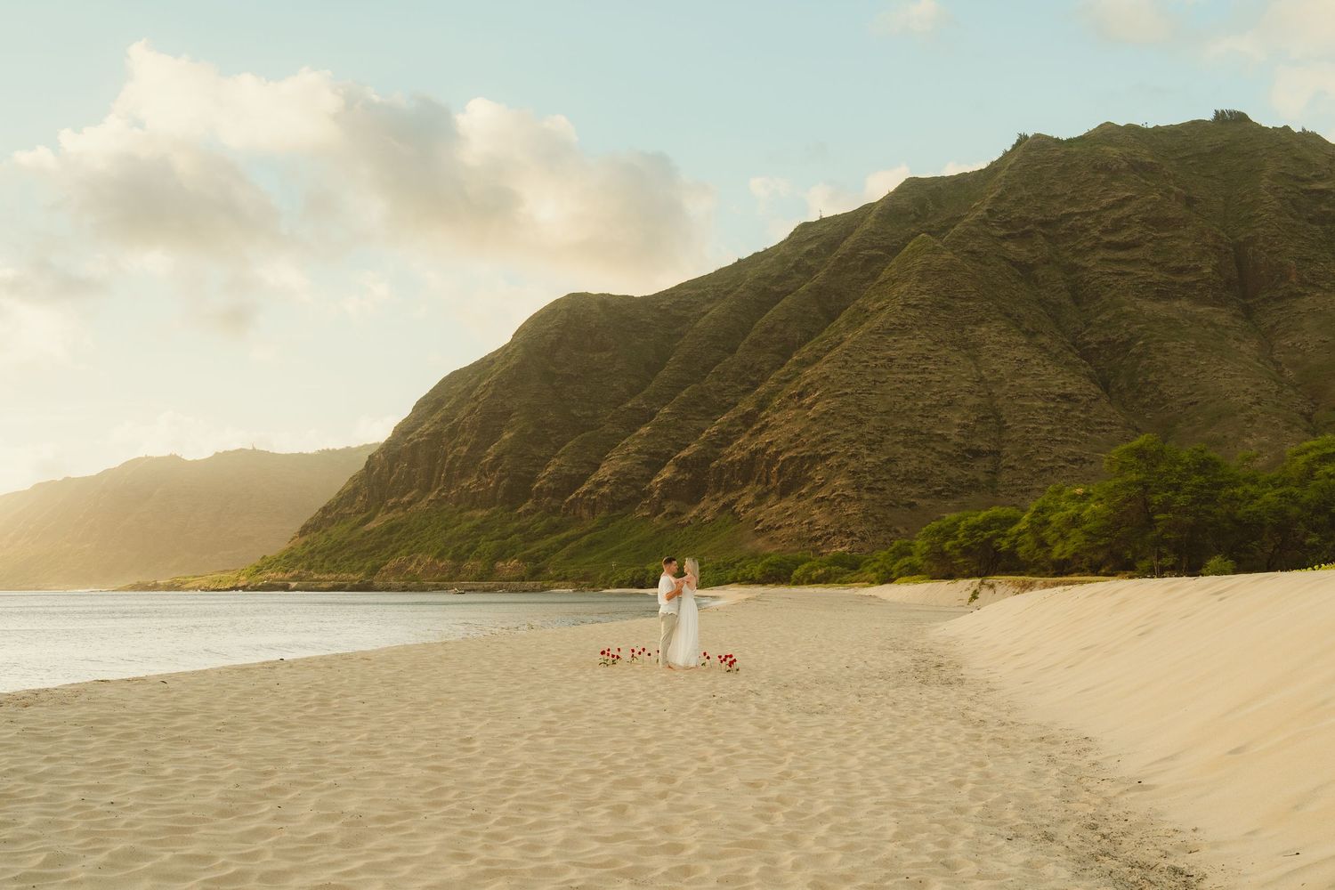 Couple in white attire stands on a sandy Hawaii beach with lush green mountains and golden sunset light behind them.