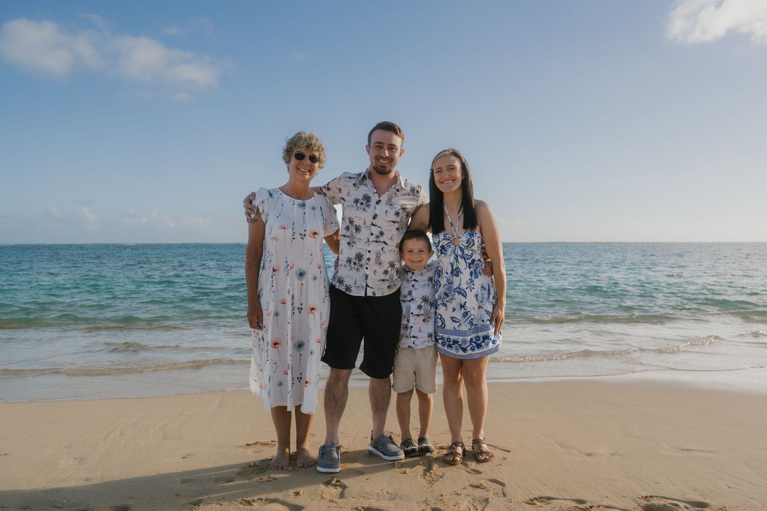 Family of four posing together on a sunny Hawaiian beach with turquoise ocean waves in the background.