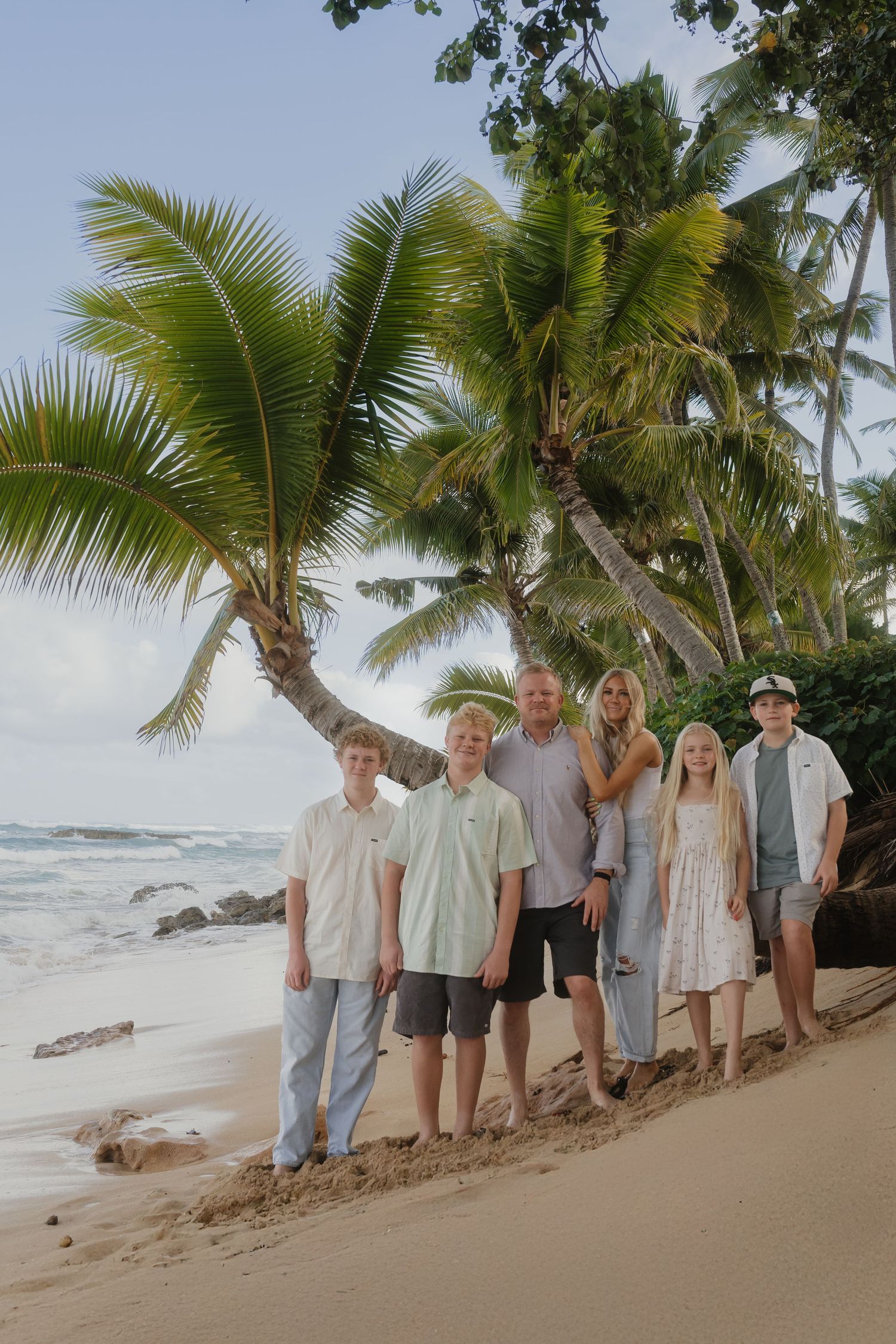 Six-member family smiling together under leaning palm tree on a sunny tropical beach.