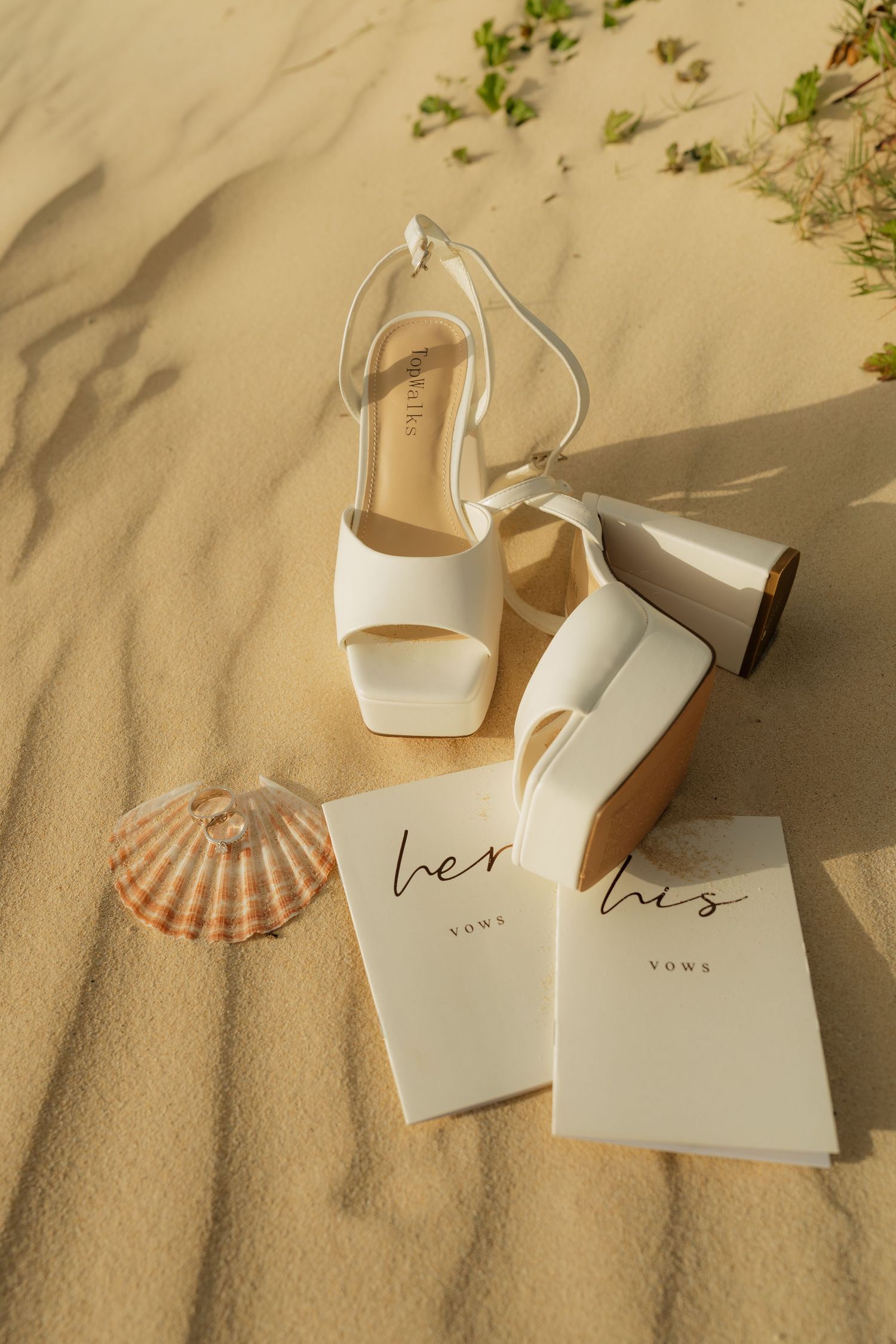 White bridal heels with vow books labeled his and hers on sandy beach with seashell.