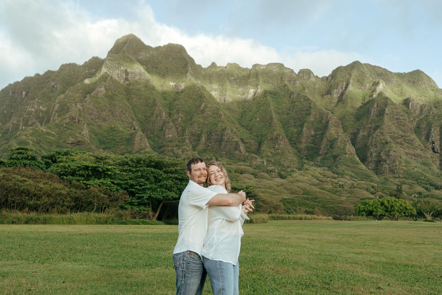 Happy couple hugging on green lawn with stunning Kualoa Ranch mountain range backdrop.