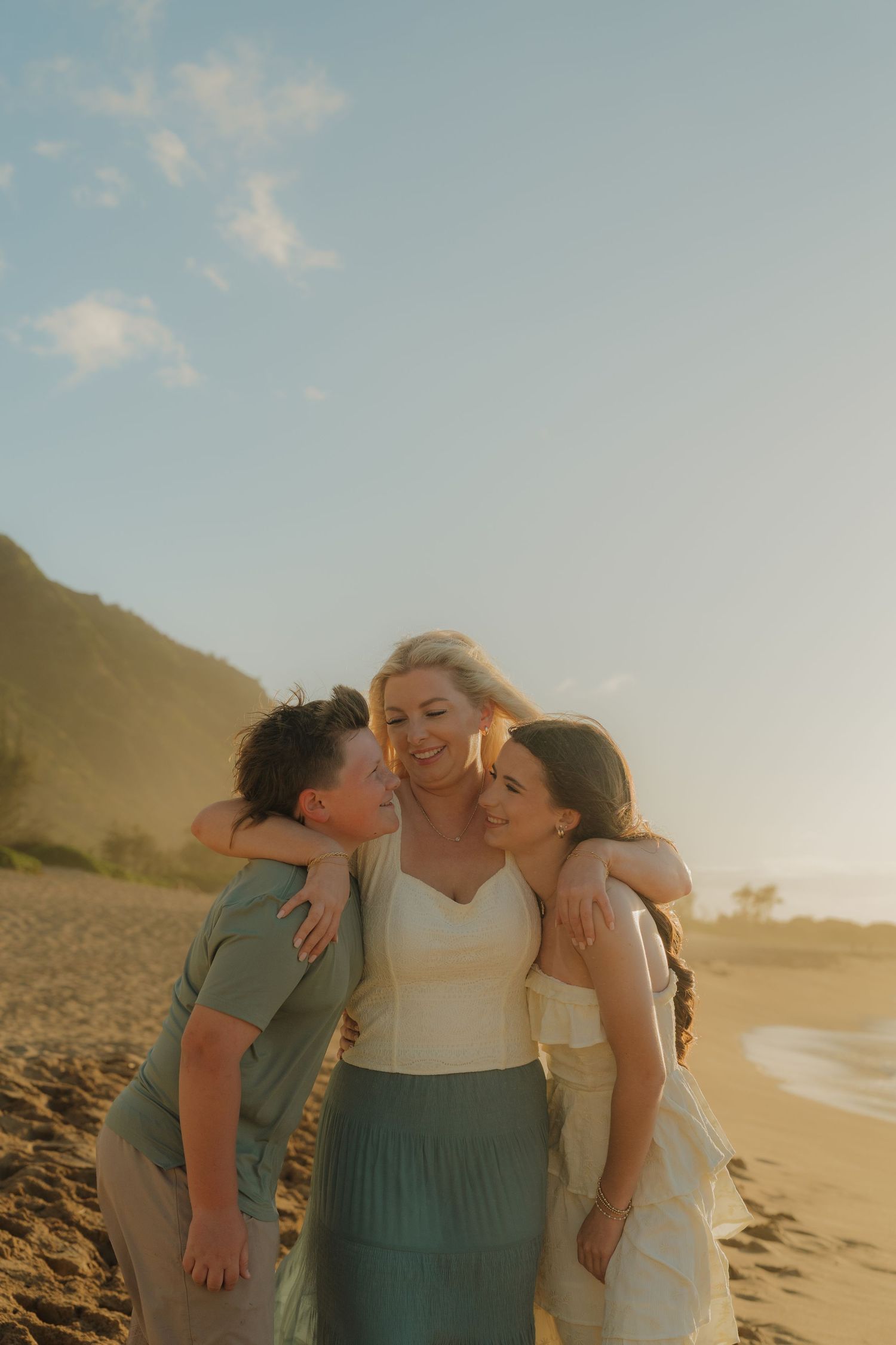 Three women embrace on a sandy beach at golden hour, smiling warmly with mountains and blue sky behind them.