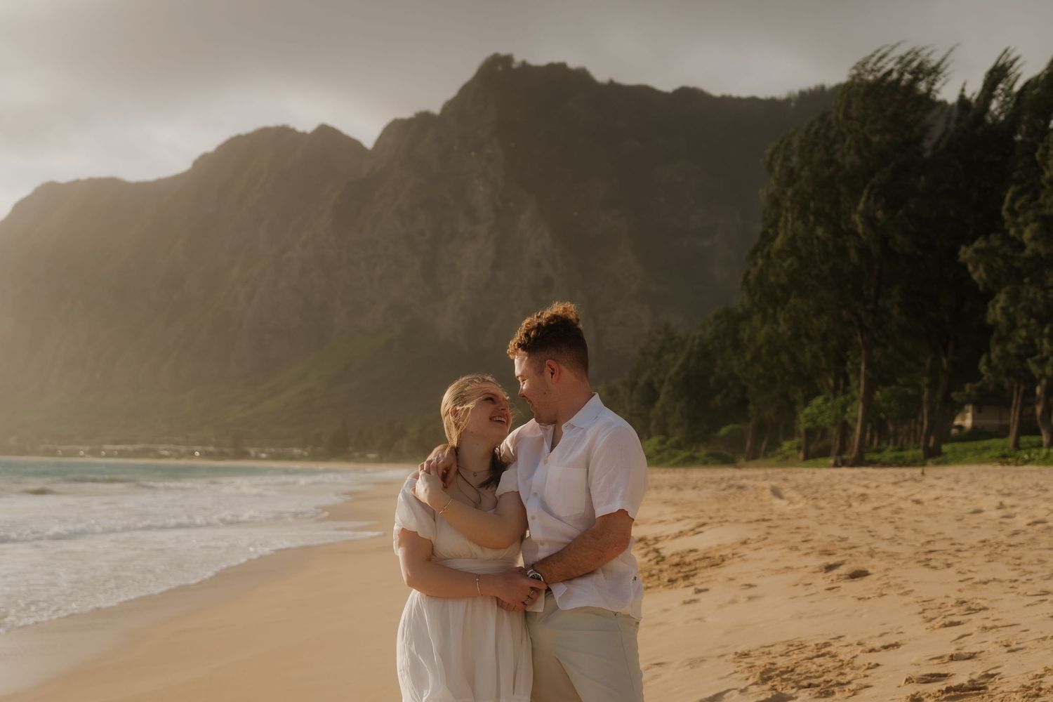 Couple embracing on a Hawaiian beach at sunset with lush green mountains in the background.
