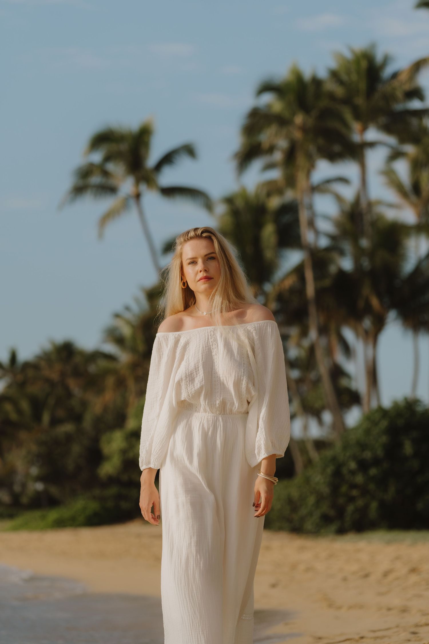Blonde woman in white off-shoulder maxi dress walking on tropical beach with palm trees.